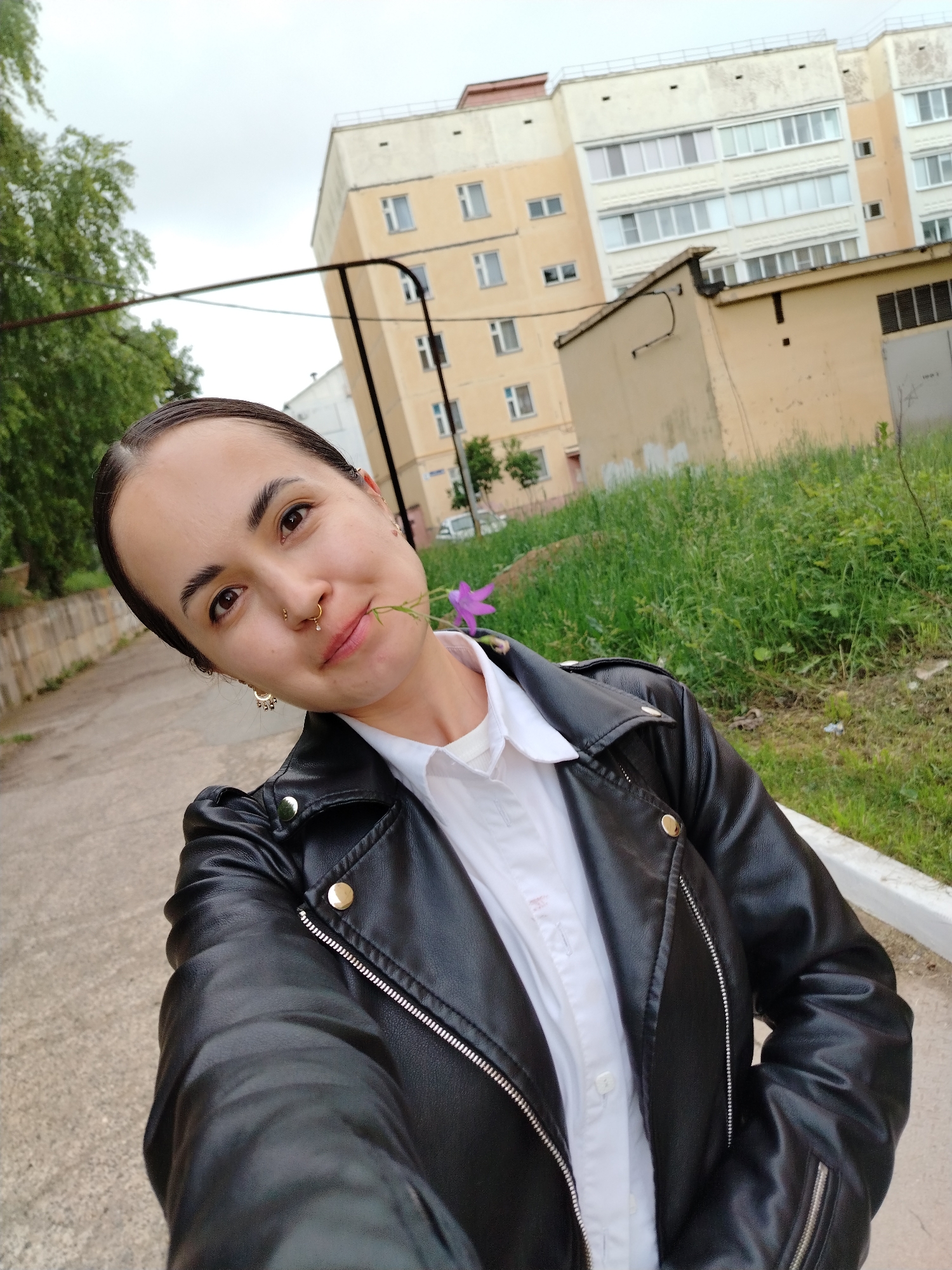 The image depicts a female individual taking a selfie in what is reportedly the city of Noviy, Russia, under an overcast sky. The subject, a young adult with dark hair pulled back, brown eyes, and thick eyebrows, looks directly at the camera. She has a gold-toned septum ring and two small stud piercings on her right nostril. Her visible left ear displays a gold-toned dangle earring. She wears a white collared shirt underneath a black leather-style jacket featuring silver zippers and gold-toned snap buttons. A single purple flower with a green stem is held between her lips, extending from her left cheek.
The background consists of an outdoor urban environment. To the left, a concrete pathway or sidewalk runs alongside a row of green trees. To the right, a patch of overgrown green grass and weeds separates the pathway from buildings. In the midground, a light yellow/beige multi-story apartment building with numerous windows and balconies is prominent, appearing slightly angled. In front of this larger structure, a smaller, single-story utility building or garage, also light yellow/beige, is visible, with a metal gate partially seen on its right. A dark horizontal wire or pipe structure, supported by a vertical pole, spans across the mid-ground in front of the buildings.