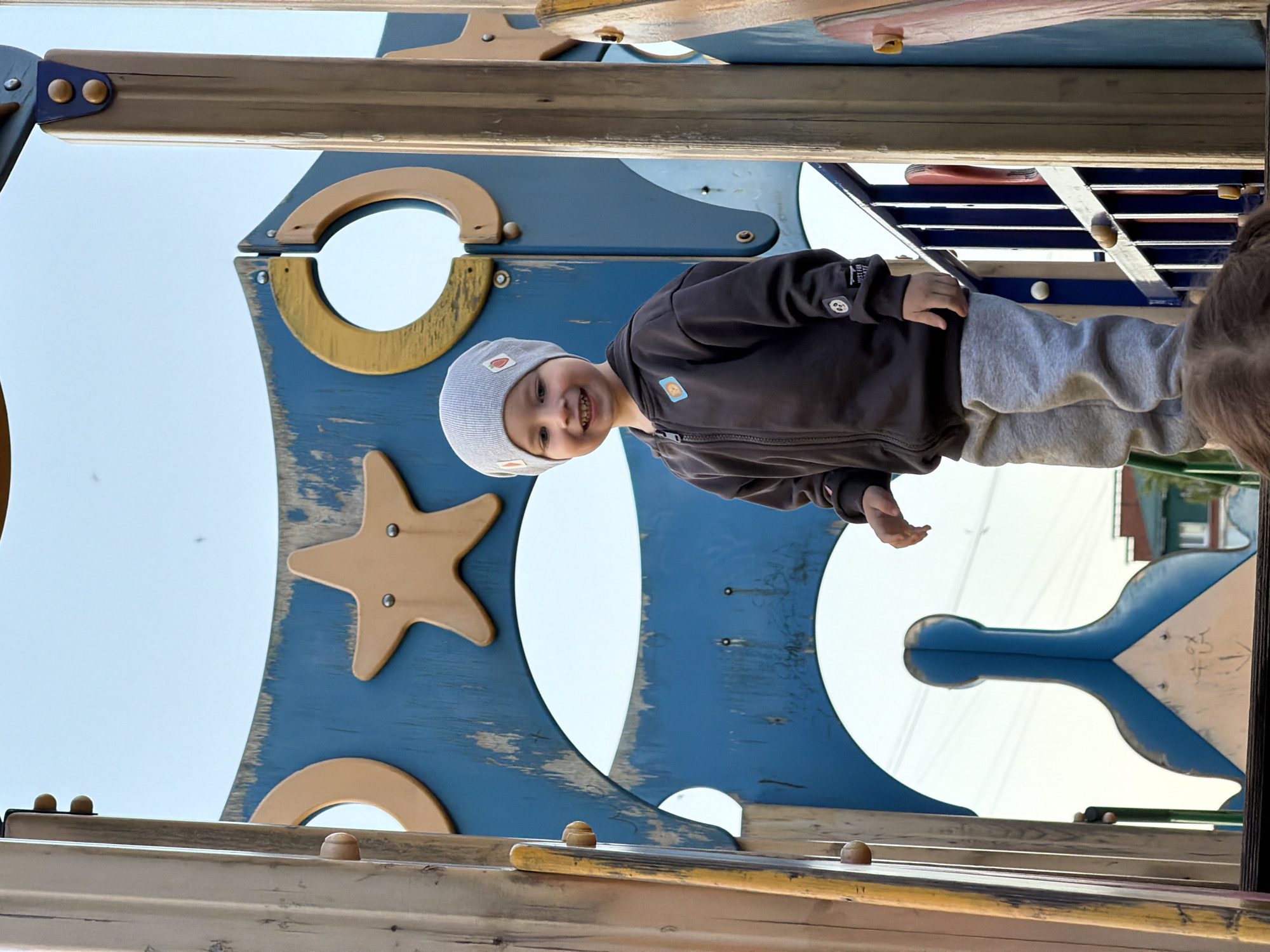 A joyful young child is captured in a vertical-oriented medium shot, smiling brightly at the camera while standing on a wooden playground structure in Chaikovsky, Russia. The image appears to have been rotated 90 degrees clockwise, placing the child on what would typically be a side panel, looking up.
The child, likely a toddler or preschooler, wears a light grey beanie adorned with small patches, including a red heart. They are dressed in a dark brown or black zippered jacket, also featuring small decorative patches (one resembling a bear or lion head on the chest, and faint text on the sleeve cuff, possibly "BUSTED"). Light grey sweatpants complete their outfit. Their wide smile and bright eyes convey happiness and engagement.
The playground structure, made predominantly of wood, is painted in varying shades of blue with areas of distressed paint revealing the natural wood underneath. It features various cutouts and shapes: large circles, a prominent five-pointed star, and abstract, organic forms, all adding to a whimsical design. Wooden beams, rails, and rungs are visible, suggesting elements for climbing or navigating the structure. The bright, clear sky forms the background, indicating daytime and good weather. In the distance, through the gaps in the structure on the right, some indistinct building shapes can be seen. Another person's head and hair are partially visible on the far right, suggesting an adult or another child nearby, possibly observing or interacting with the main subject.