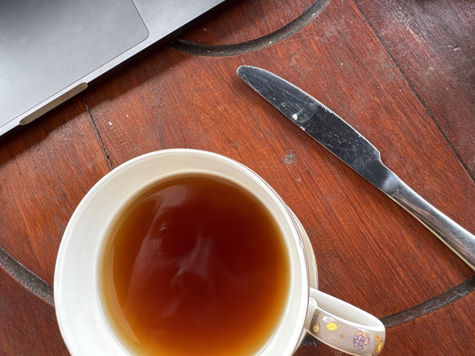 An overhead, close-up shot captures a casual scene on a reddish-brown wooden table. The primary subjects are a white ceramic cup filled with a dark amber liquid, a silver laptop, and a metal butter knife.
The cup, positioned prominently in the lower left, contains what appears to be tea or coffee. A distinct reflection of a human face looking down into the drink is visible on its surface. The cup's handle, seen on the right, features decorative elements with abstract yellow, purple, and gold patterns.
In the upper left quadrant, the edge of an open silver laptop is visible, showing a small rectangular slot on its side. A dark cable extends from near the laptop, crossing over the table surface. To the right of the cup and partially under the laptop, a well-used metal butter knife with a serrated edge rests diagonally on the table. The knife's blade shows visible scratches and wear.
The wooden table has a rich, warm tone, displaying prominent grain, a visible crack running across its surface, and subtle circular patterns that could be part of its design or wear marks. Scattered specks of dirt or dust are also visible on the table and the knife. The scene suggests a relaxed moment of working or leisure. No text is visible in the image.