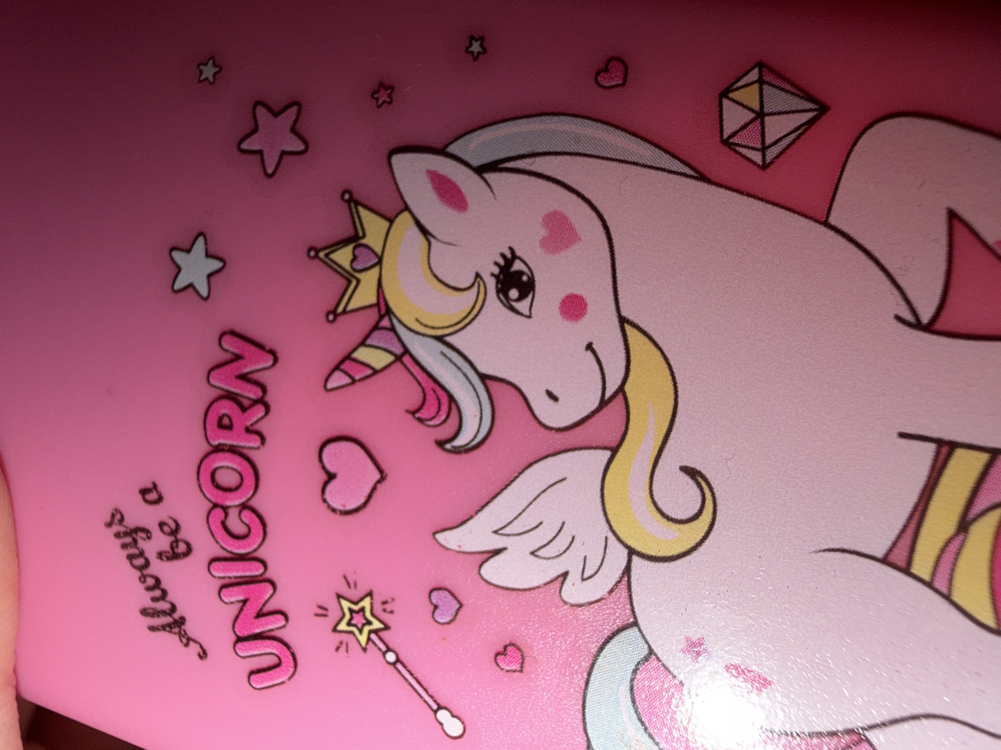 The image displays a pink surface with a drawing of a unicorn. The unicorn is light pink with a yellow mane and tail, a yellow crown with a pink heart on it, and a striped horn. It has a white wing on its back. There are several smaller illustrations on the pink surface, including a magic wand, hearts, stars, and a diamond. The text "Always be a UNICORN" is printed above the unicorn. There is no indication of a location in Varna, Bulgaria.