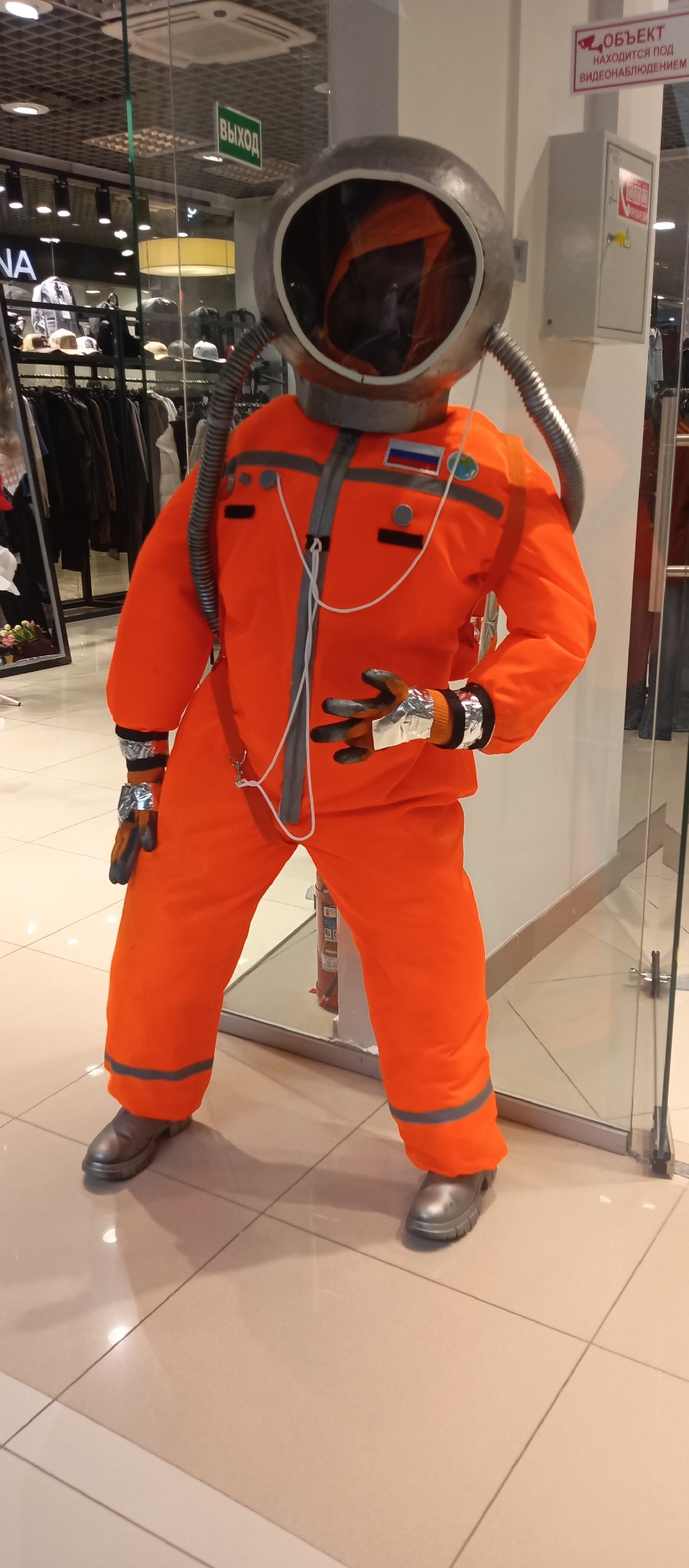 An orange astronaut suit is on display inside a shopping mall in the city of Unknown. The spacesuit has a large grey helmet and two silver tubes. The suit has gray and silver colored gloves and shoes. A Russian flag is visible on the left shoulder. The tiles are beige with brown grout. A clear glass wall runs along the right. A fire extinguisher is partially visible behind the spacesuit. A sign in Russian that translates to “Object under video surveillance” is visible above the astronaut on the right.
