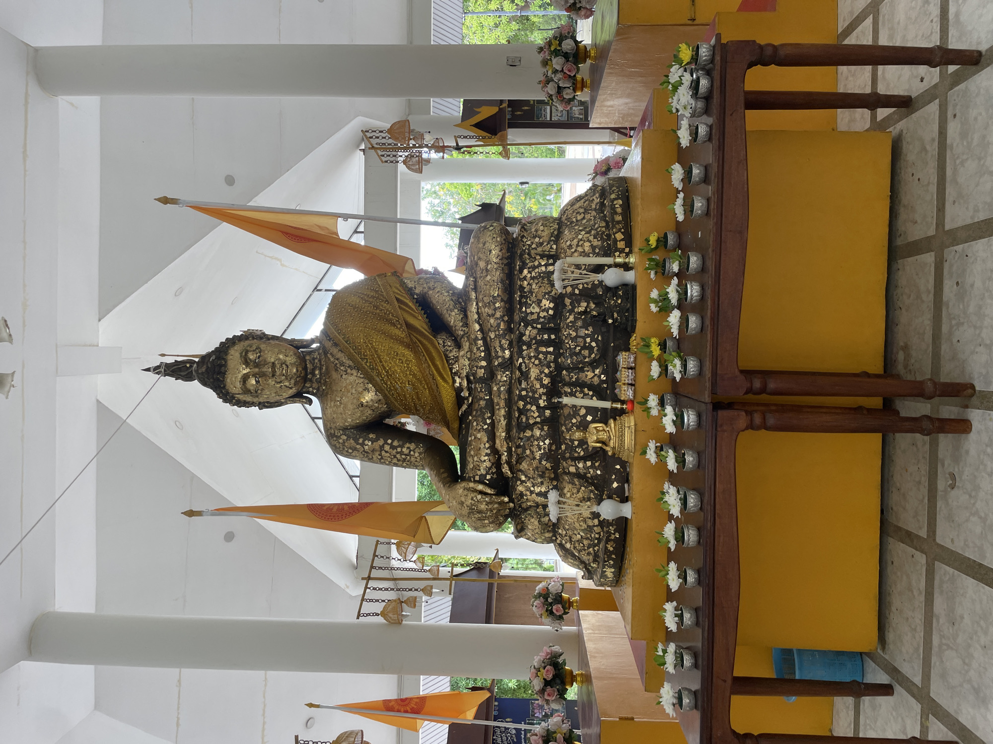 In Na Chom Thian, Thailand, a large, seated golden Buddha statue is the central focus of this image. The statue is adorned with gold leaf, suggesting it's a significant religious monument. It sits on a raised platform, also covered in yellow, with a smaller golden Buddha statue placed in front of it. Surrounding the main statue are offerings: small white and yellow flowers in silver pots arranged on two wooden tables in the foreground, a white vase with incense sticks, and a lit candle.
Two large orange flags with Buddhist symbols are positioned behind the main Buddha, flanking it. The structure itself appears to be an open-air pavilion or temple hall, indicated by white pillars supporting a white ceiling with visible beams and a sloped roof structure. Hanging from chains above are decorative bamboo-like structures with bells. In the background, glimpses of green trees and foliage suggest an outdoor setting beyond the pavilion. The floor is tiled in a pattern of light and dark grey squares. A blue recycling bin is visible to the left, behind the wooden table.