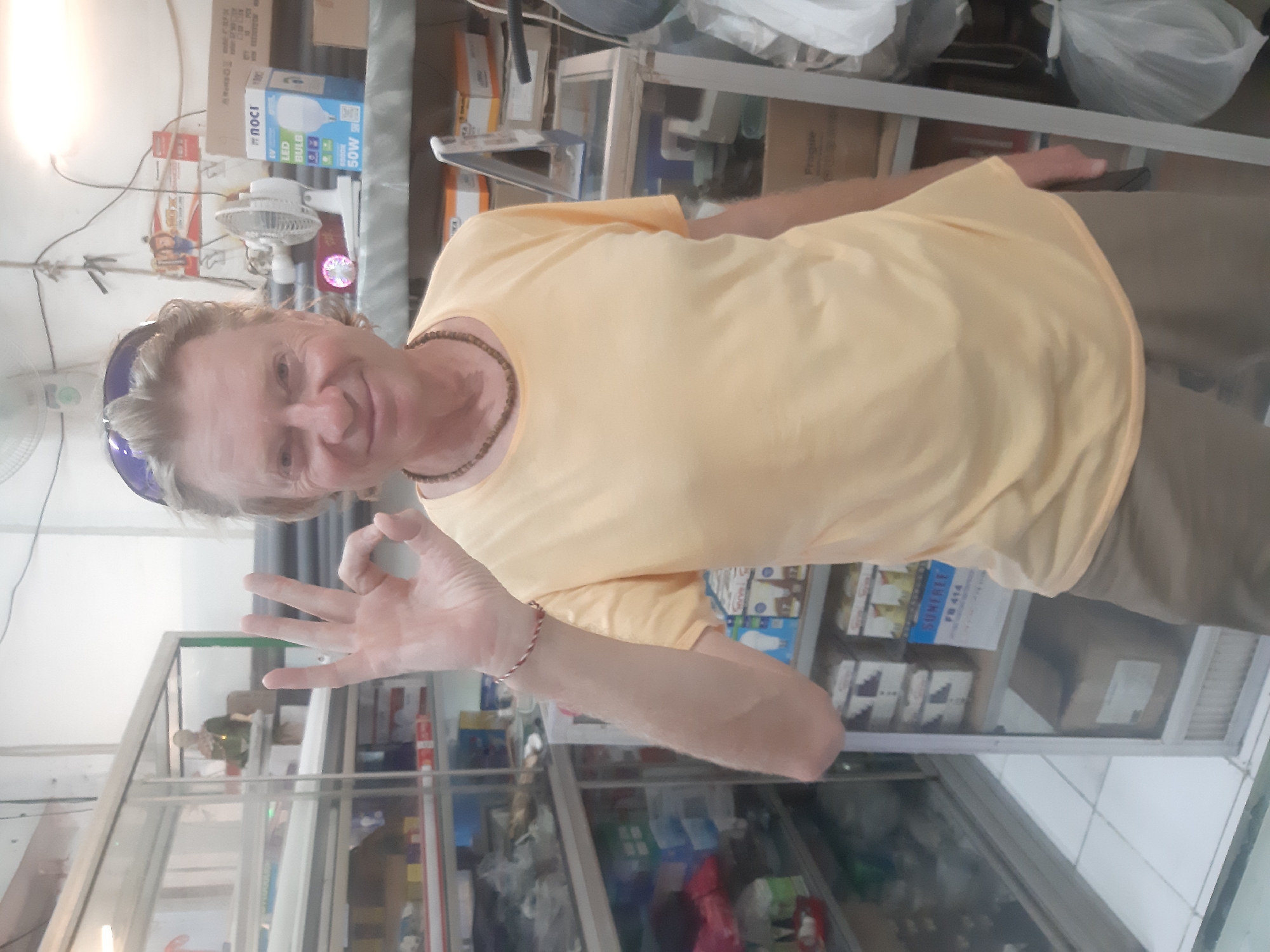 A man with gray hair and blue sunglasses on his head is standing in what appears to be a shop in Denpasar, Indonesia. He is wearing a yellow t-shirt and khaki pants. He is making an "OK" hand gesture with his right hand. Behind him is a glass display case filled with various items. Above the display case are shelves with more items, including a small white fan and boxes of light bulbs. The background is cluttered with boxes and other objects, suggesting a store or storage area. The floor is tiled.