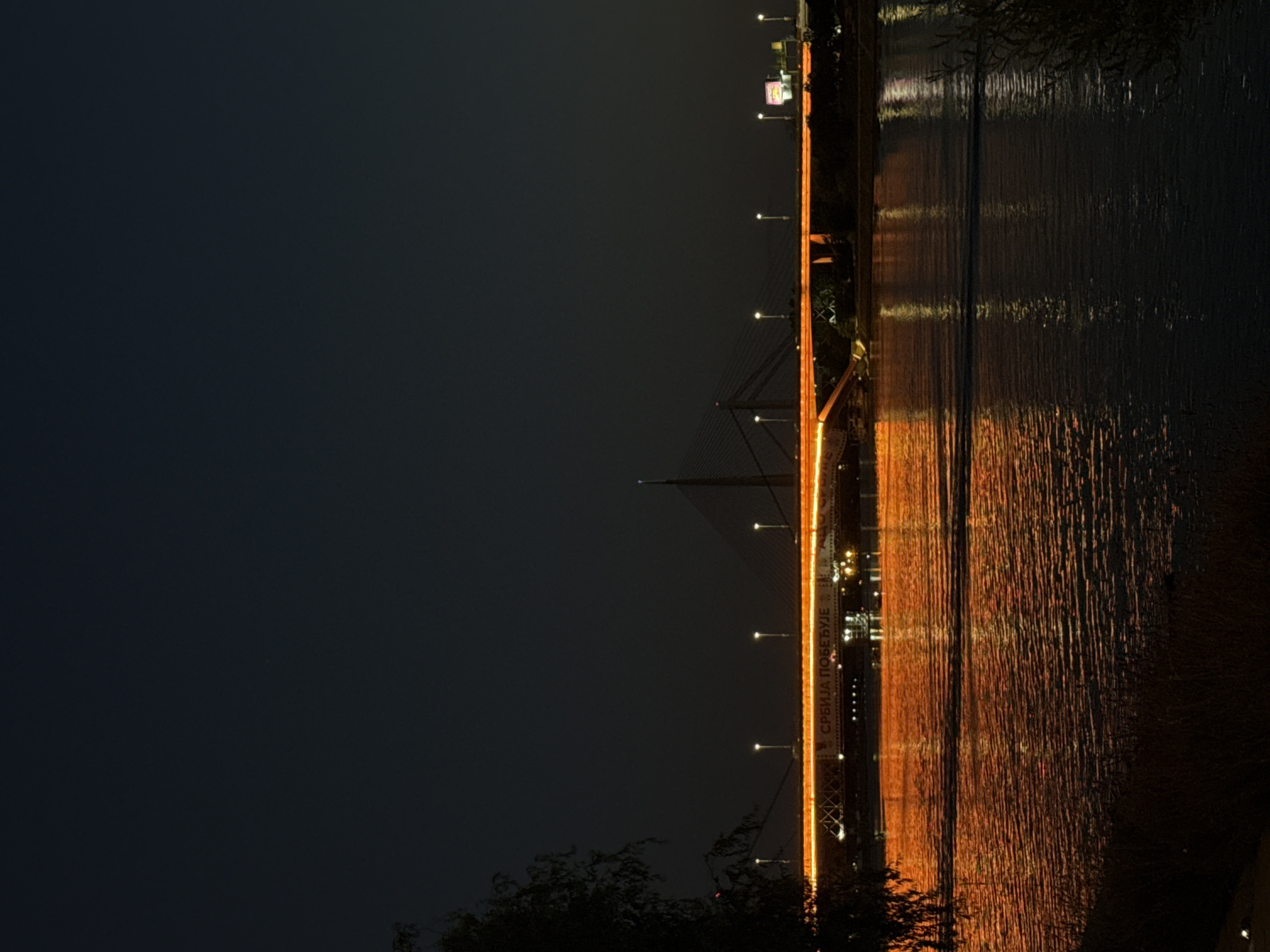 The image is a long shot of the Ada Bridge at night in Belgrade, Serbia. The bridge is illuminated with orange lights, and its reflection is visible in the dark water of the Sava River below. Several lampposts are also lit along the bridge, casting a warm glow. In the foreground, the dark silhouettes of trees and vegetation are visible on the riverbank. The sky is dark, suggesting it is nighttime, with a few distant lights from the city. There are no people visible in the image. The overall atmosphere is serene and picturesque.