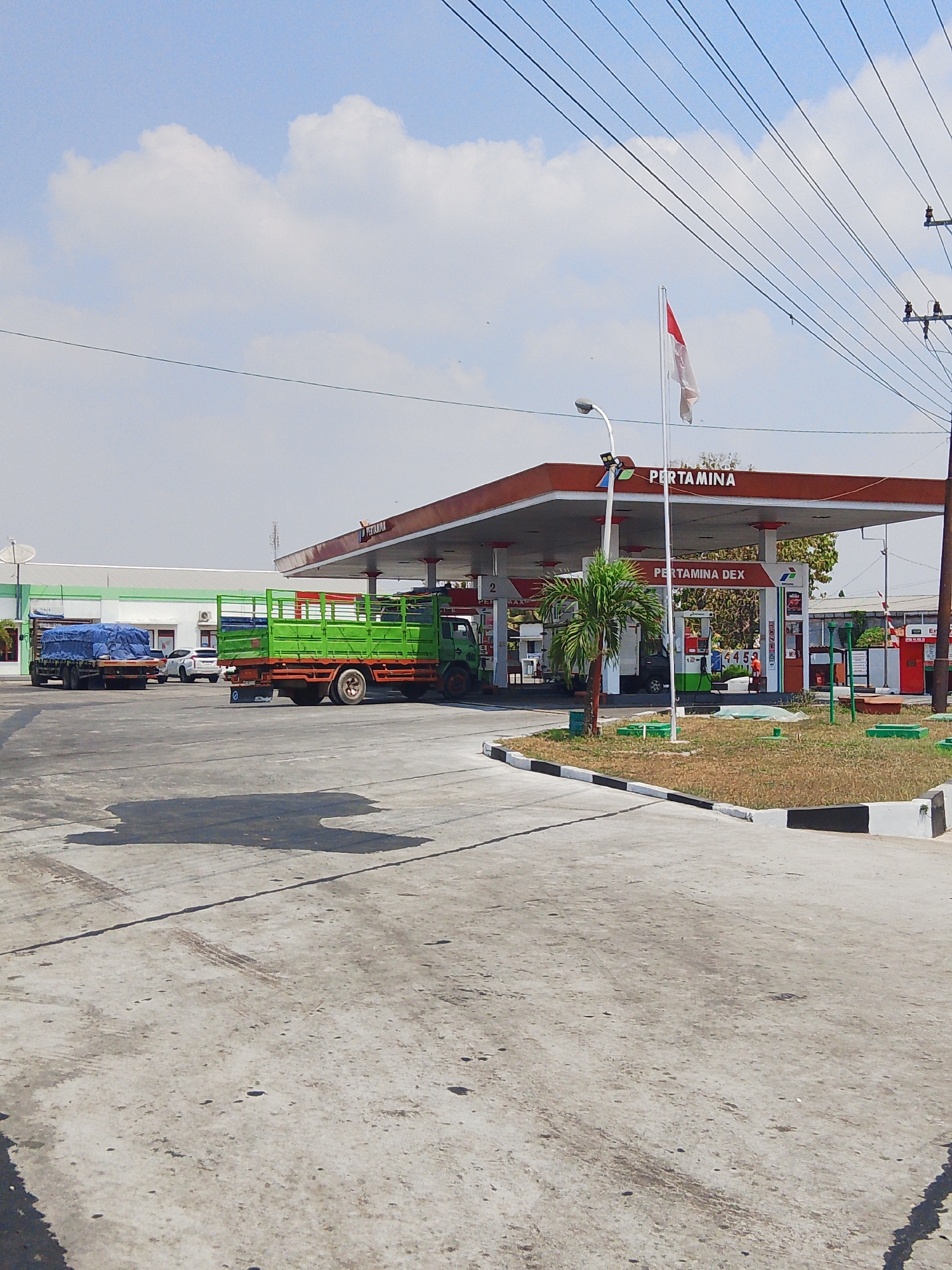 The image depicts a daytime scene at a Pertamina gas station in Sedo Lor, Indonesia. The gas station features a red and white awning, supported by white pillars. The Pertamina logo is prominently displayed on the awning. Several vehicles are present, including two trucks. One truck, partially covered with a blue tarp, is parked on the left side of the image. A bright green truck with an orange chassis is positioned closer to the gas station, appearing to be actively serviced or parked. A white SUV is visible behind the tarp-covered truck. The ground surface is a mix of concrete and asphalt, with some dark stains and tire marks visible. In the background, there are buildings with green and white facades, and utility poles with overhead wires. A flagpole with the Indonesian flag (red and white) is positioned near the gas station. A small palm tree stands to the right of the main gas pumps. The sky is blue with scattered white clouds. The environment appears to be an open, somewhat industrial or commercial area. There are no identifiable people in the image.