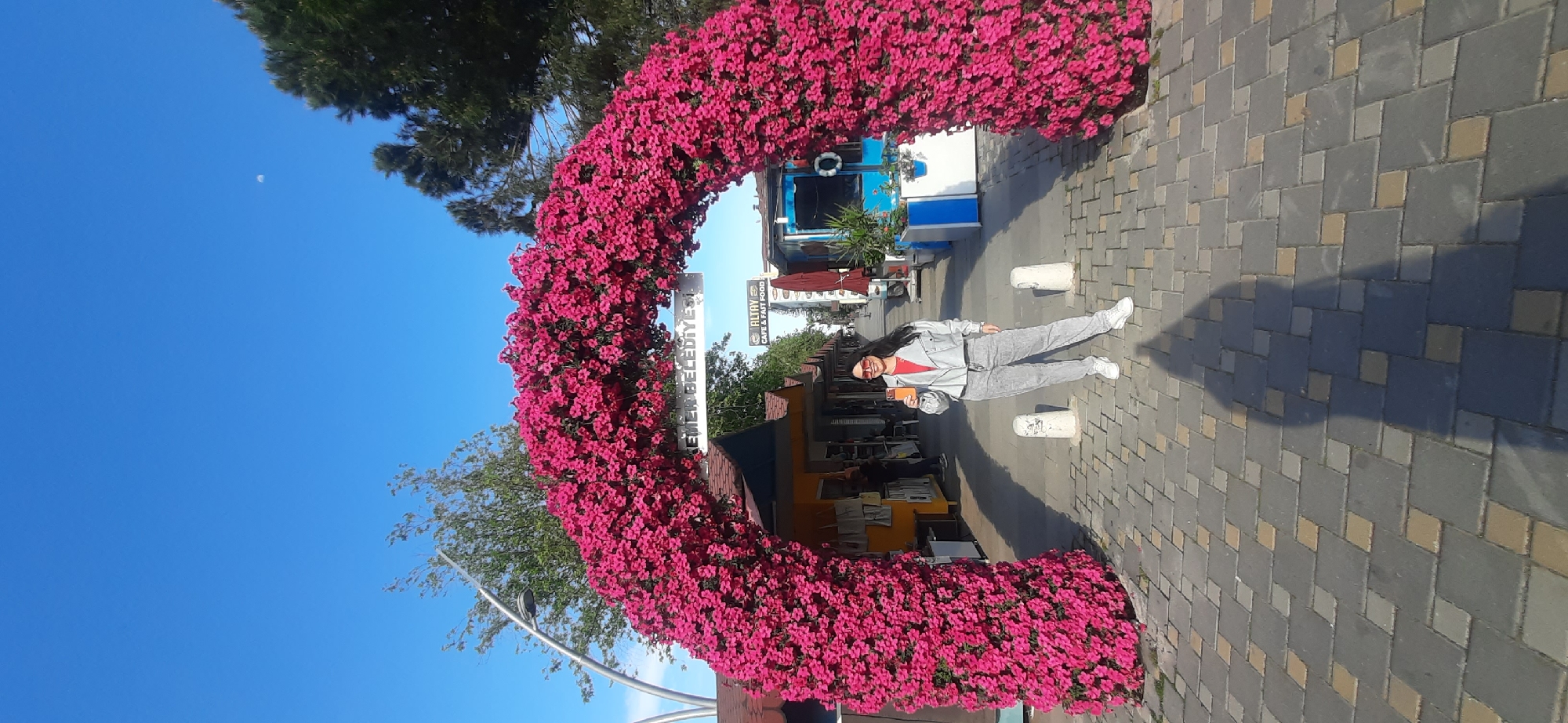 A vertically oriented outdoor photograph captures a woman posing in front of a floral archway on a paved surface. The archway is densely covered with vibrant pink flowers, forming a semi-circular shape. Beyond the archway, there are commercial establishments, including a cafe called "Altay Cafe & Fast Food," and street signs indicating the location as Kemer Belediyesi, Turkey.
A woman stands to the right of the archway. She wears a light gray tracksuit, white sneakers, and sunglasses. She holds an orange object in her hand. Her shadow stretches on the paved surface, which is composed of gray and brown rectangular blocks.
The sky in the background is clear and blue. Trees are partially visible on the left side of the frame, near the archway. A small, faint crescent moon is visible in the upper left corner of the sky.