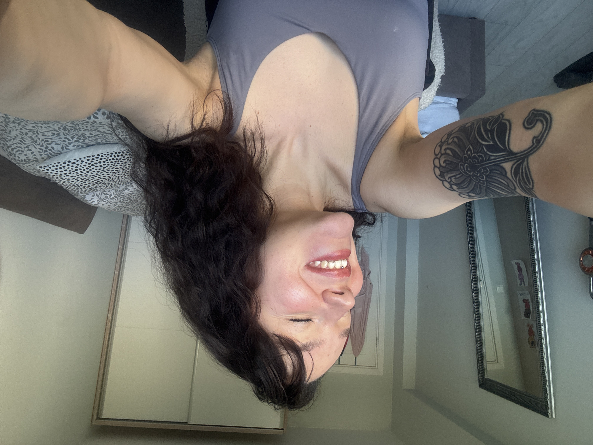 A woman with dark wavy hair, fair skin, and a tattoo on her right upper arm is smiling with her eyes closed. She's wearing a gray tank top. The background includes a bed with patterned pillows, a gray couch, and a hallway with a closet. A mirror with a decorative frame is visible on the wall. The exact location within Burgas, Bulgaria cannot be determined from the image alone.