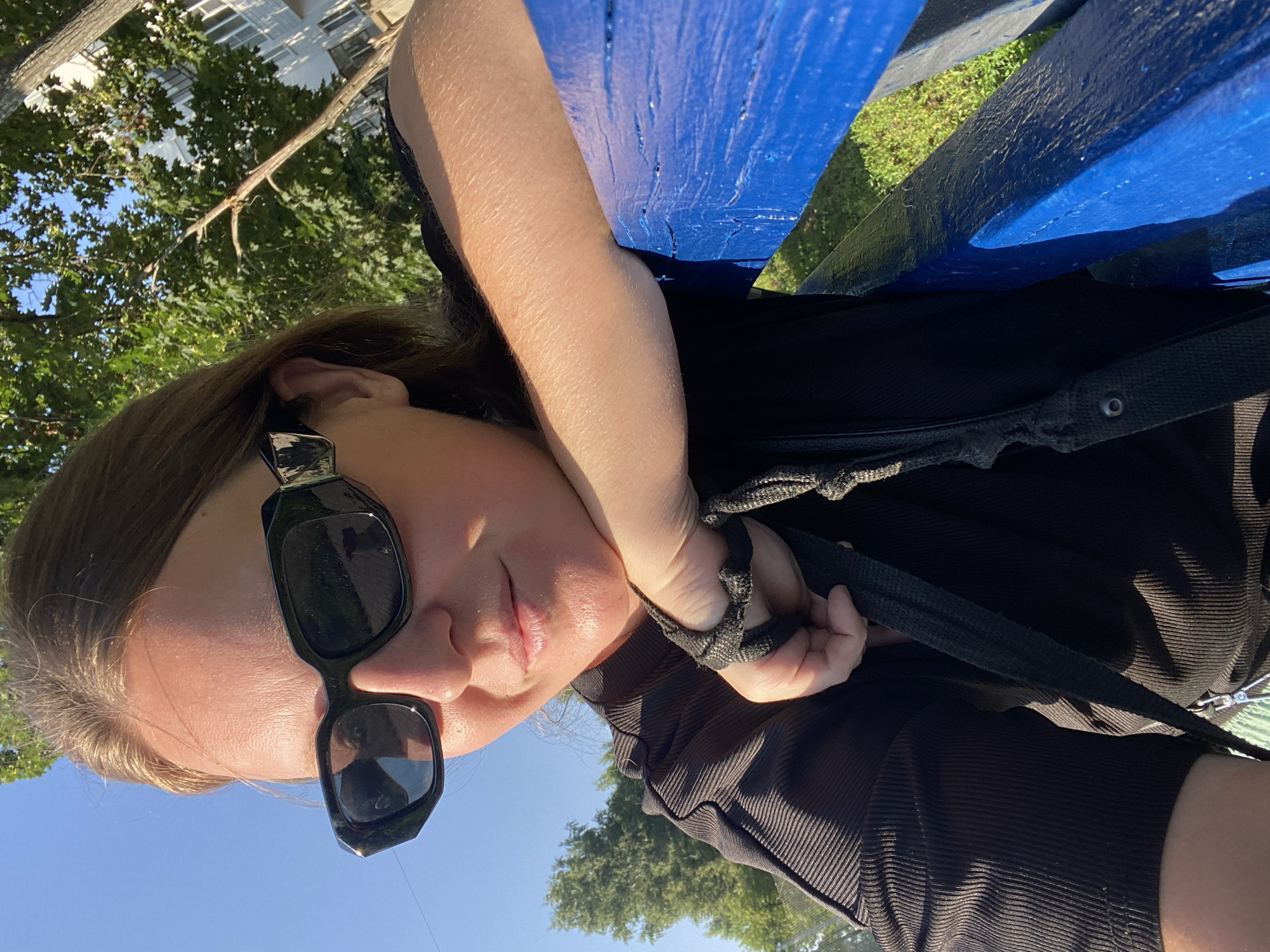 A woman wearing black sunglasses and a black shirt poses for a selfie outdoors on a sunny day. She leans on a bright blue bench, with her right arm resting on it and her hand supporting her chin. Her left hand is visible, holding onto a black strap. The background shows green trees and a building, suggesting an urban park or residential area. The lighting indicates it is late afternoon.