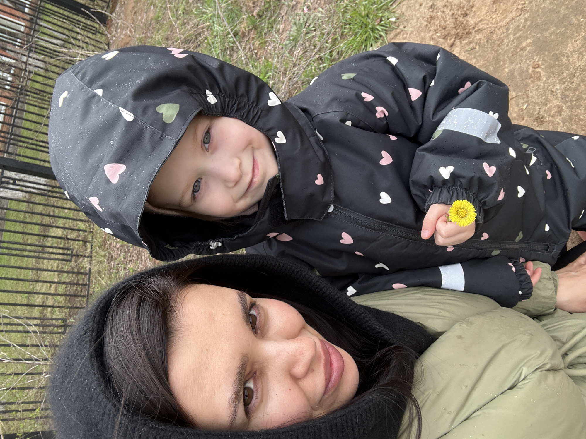 A woman and a child are lying on the ground. The child, wearing a black hooded jacket with heart patterns, holds a yellow flower in their hand. The woman, with dark hair, is also wearing a hood. The background suggests an outdoor environment, possibly a park or yard. Based on available information, the location is Chaikovsky, Russia.