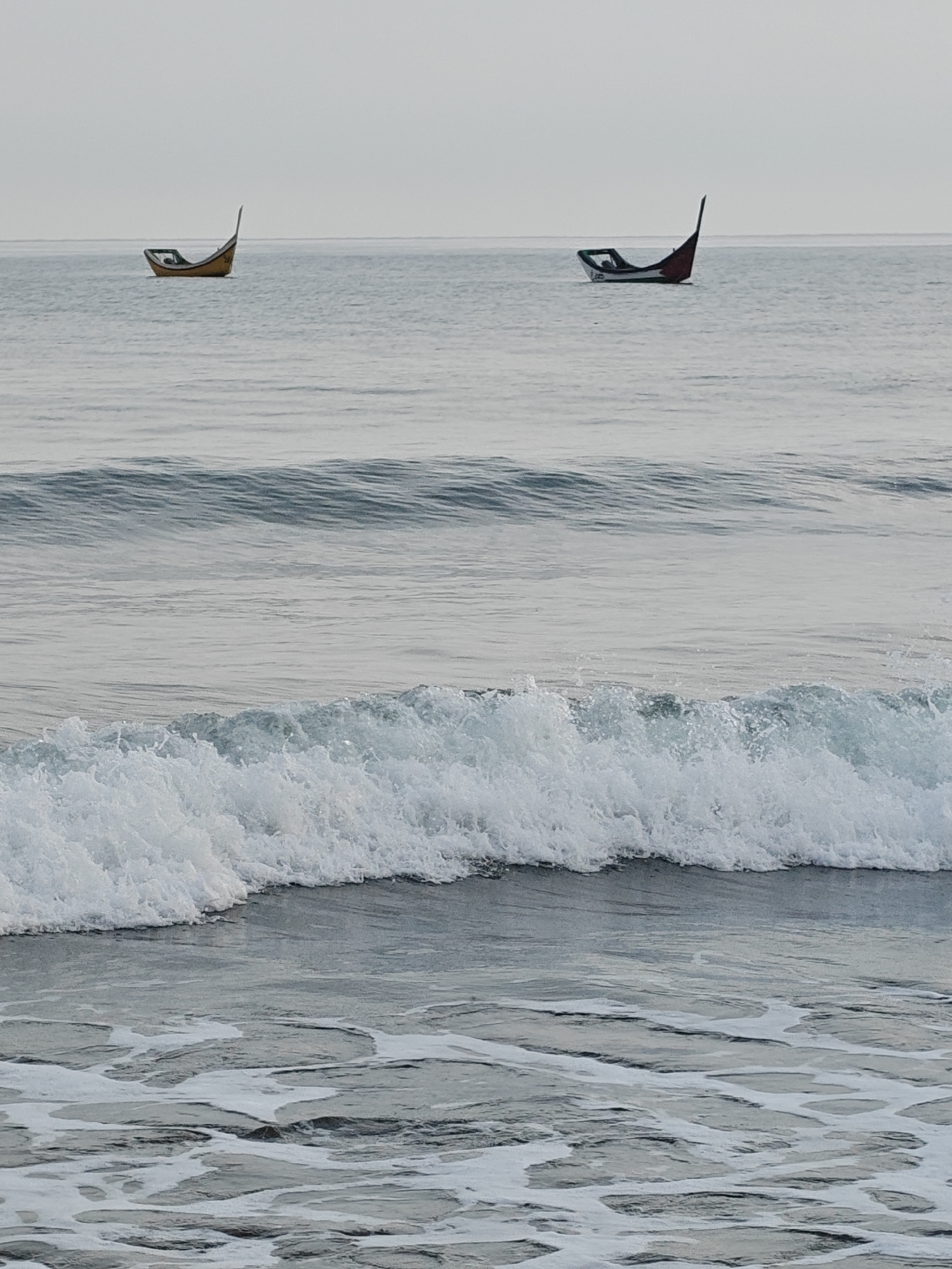 Two small, empty fishing boats are adrift on a calm sea under an overcast sky. The boat on the left is yellow with a white interior and a tall, pointed stern. The boat on the right is white with red and dark green accents on its stern, which is also tall and pointed. The water is a muted grey-blue, with gentle waves and some white foam in the foreground. The horizon is indistinct, blending the sea and sky. The scene is devoid of human presence and suggests a quiet, possibly early morning or late afternoon setting. The location is identified as Curehtunong, Indonesia.