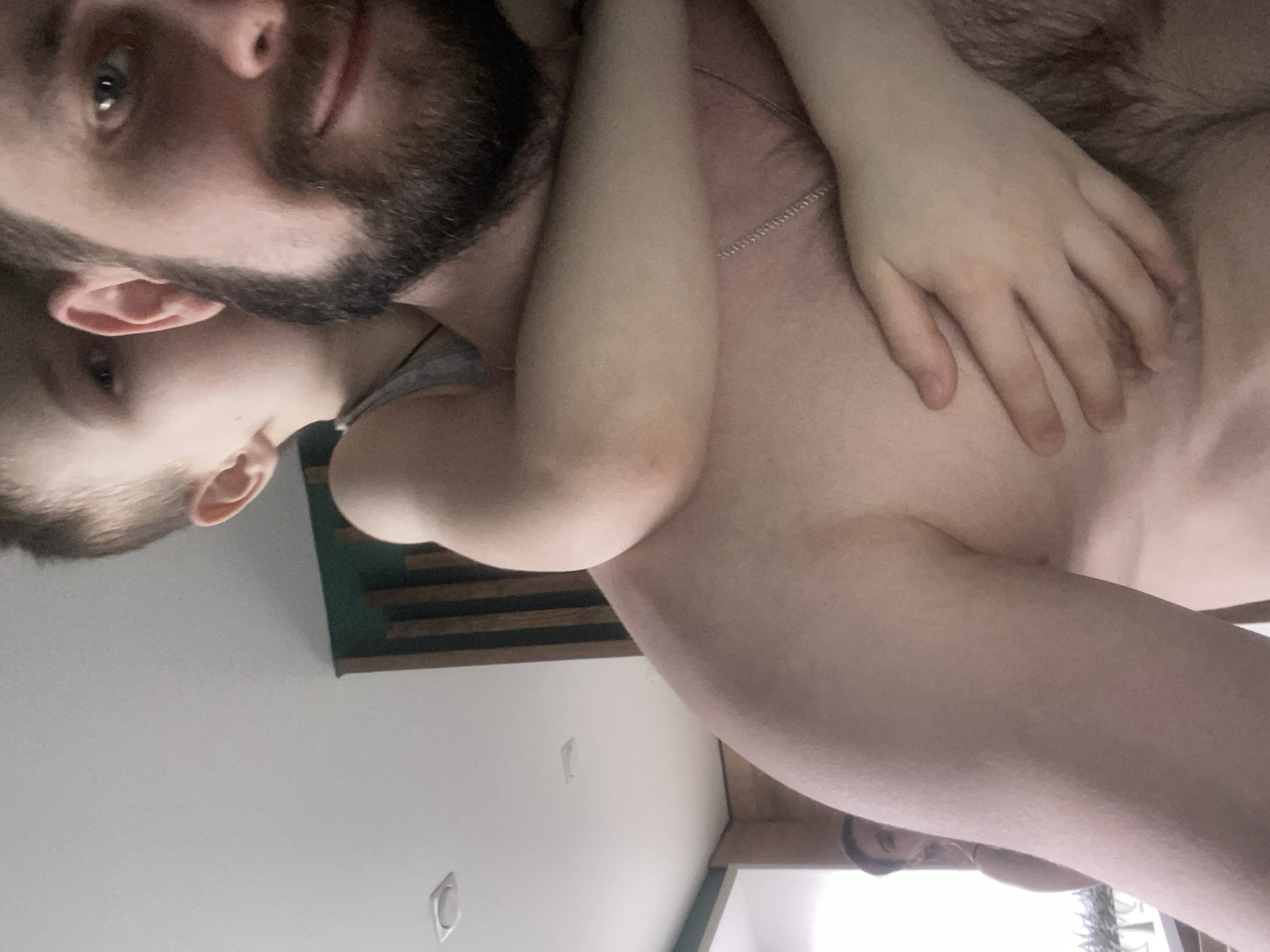 A man with a beard is lying down, holding a child. The man is facing the camera, and the child's head is near his ear. The child's hand is on the man's chest. A silver chain is around the man's neck. The location is possibly an interior with a green and brown decorative panel on the wall and a white ceiling with light fixtures. The location is unknown and there is no evidence it is in Kazan, Russia.
