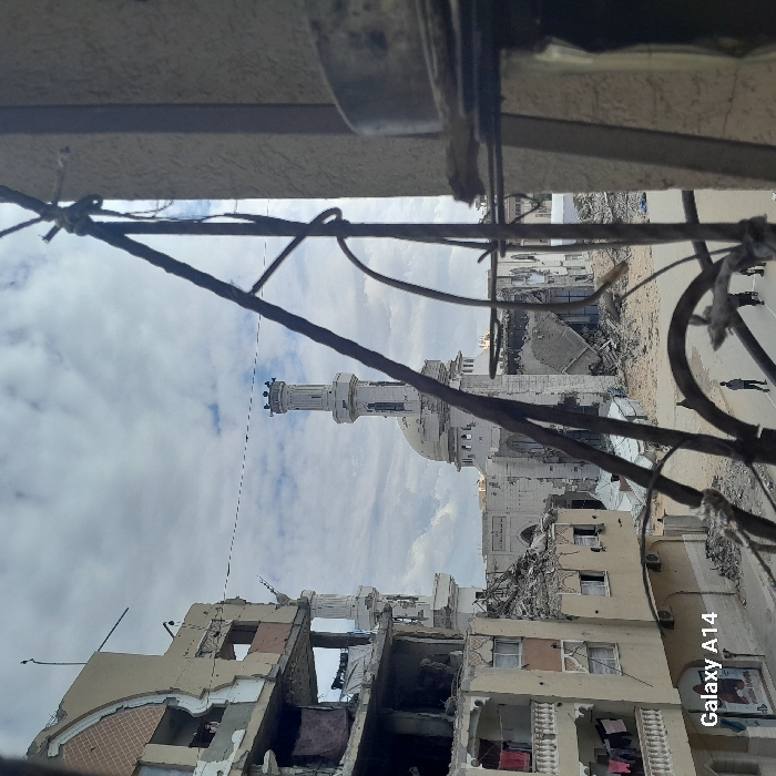This image depicts a scene of extensive urban destruction in Khan Yunis, Palestinian Territory, under an overcast sky with scattered clouds. In the mid-ground, a large white mosque with a prominent dome and a tall minaret stands, exhibiting visible damage but largely retaining its structural integrity. To the left of the mosque, a multi-story building, likely residential, is severely damaged, with multiple floors partially collapsed, exposed interiors revealing debris, and large sections of its facade missing. Below this building, on the left, another partially damaged structure shows several air conditioning units and clothes hanging from lines, suggesting continued or recent human presence.
The foreground is partially obscured by dark, rusty, bent metal bars and wires, possibly part of a window frame or balcony railing, with a vertical concrete column segment visible on the far right. In the lower-mid ground, a paved street runs horizontally, on which at least four individuals, appearing as adult males, are walking. The ground around the damaged buildings and the street is littered with rubble, concrete debris, and twisted metal. The overall environment conveys significant infrastructural damage and widespread demolition. The "Galaxy A14" watermark is visible in the bottom-left corner.