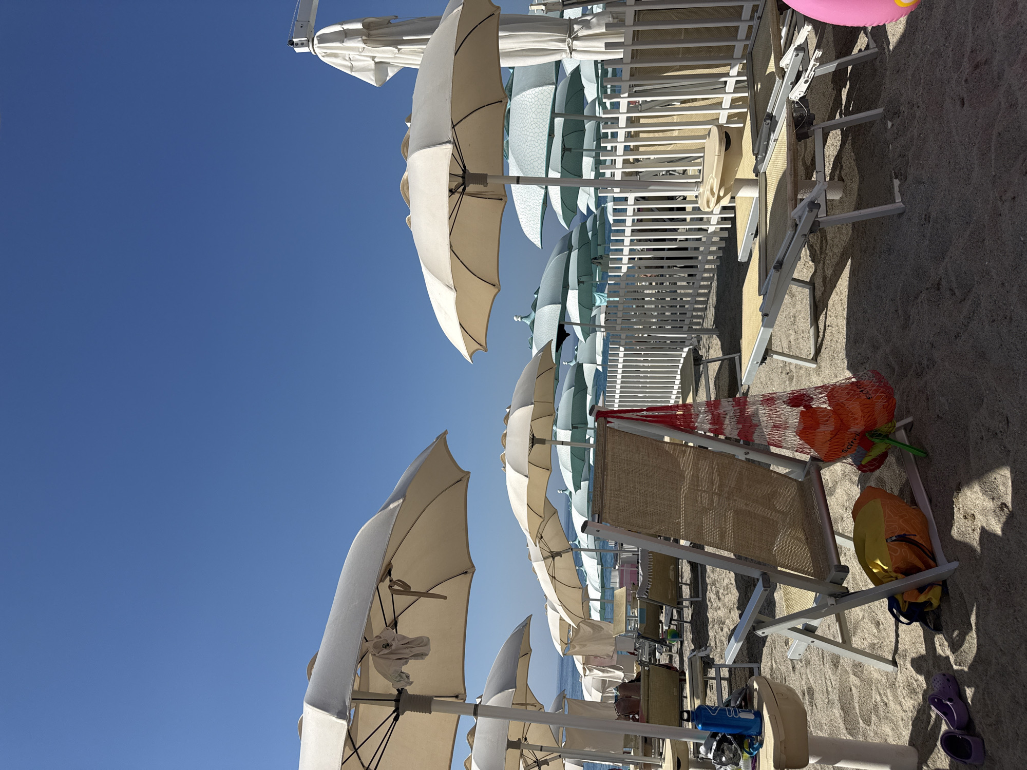 This image depicts a beach scene in Finale Ligure, Italy. The foreground is dominated by a row of beach chairs and umbrellas, with the sand showing footprints and shadows from the umbrellas. To the left, a beige table holds a blue water bottle. In the center, a beige fabric beach chair is prominently displayed, with a yellow and orange bag underneath it and a red mesh bag filled with toys hanging from its side. To the right, two empty lounge chairs are set up. In the background, more umbrellas, lounge chairs, and a white picket fence can be seen. The sea is visible in the distance under a clear blue sky. A person is partially visible sitting on a lounge chair in the mid-ground, to the left.