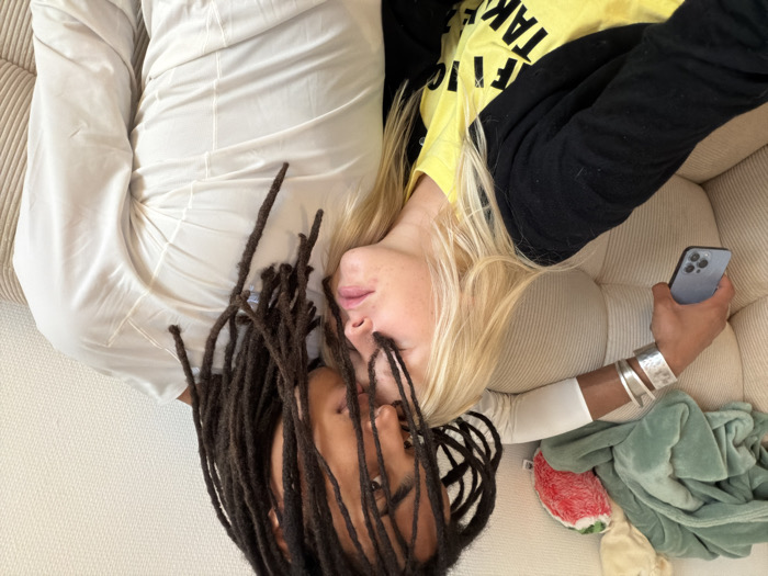 Two individuals are seated closely on a light beige, ribbed upholstered couch in an indoor setting in Paris, France. A female with blonde hair, wearing a black cardigan over a yellow t-shirt, rests her head on the chest of a male. The male, with dark skin and long dark dreadlocks partially obscuring his face, wears a light-colored long-sleeved shirt and looks down at the female. His left arm is visible, adorned with multiple silver-toned bangles, holding a dark-colored smartphone. Behind the female's head, a light green blanket and a red and white strawberry-shaped plush toy are visible on the couch. The background consists of a plain, light-colored wall.