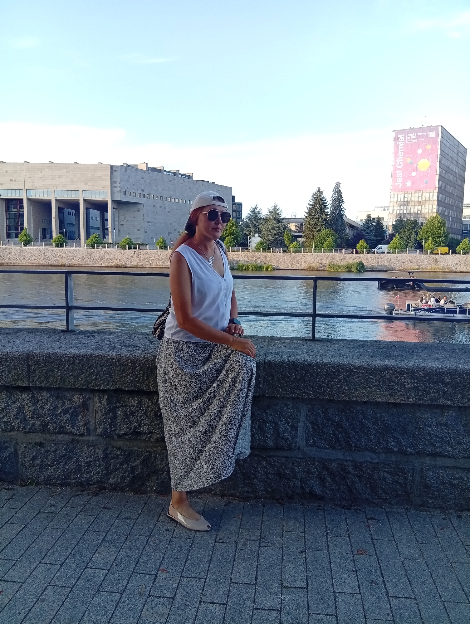A woman with auburn hair and sunglasses sits on a stone wall beside a river in Wrocław, Poland. She is wearing a white sleeveless top, a long patterned skirt, a light-colored baseball cap, and beige flat shoes. A dark crossbody bag hangs over her shoulder. In the background, there is a body of water with a boat on it. Across the river, there are modern buildings, trees, and a tall building with a large banner displaying text and graphics. The sky is clear and blue.