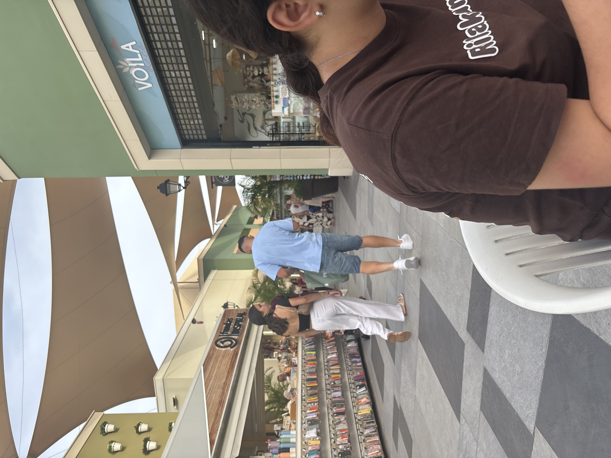 The image depicts an outdoor shopping area. In the foreground, a person wearing a brown t-shirt is seated, facing away from the camera. Behind them, a couple is walking away from the camera. The woman is wearing white pants and a black top, and the man is wearing blue shorts and a light blue t-shirt. They are walking on a paved walkway with a geometric pattern of gray and black tiles.
To the left of the couple, there is a retail stall displaying numerous items, possibly phone cases, on shelves. The stall has a wooden facade with signage that appears to read "Fundas Inspíral". Further to the left, a building with green accents is visible, featuring hanging planters with white flowers.
To the right, a building with a green facade has a storefront with a sign that says "VOILA". The storefront has large windows and a rolling metal security gate. The overall environment suggests a modern outdoor shopping mall or pedestrian street. The question of this location being in Orihuela, Spain is not directly verifiable from the visual information provided.