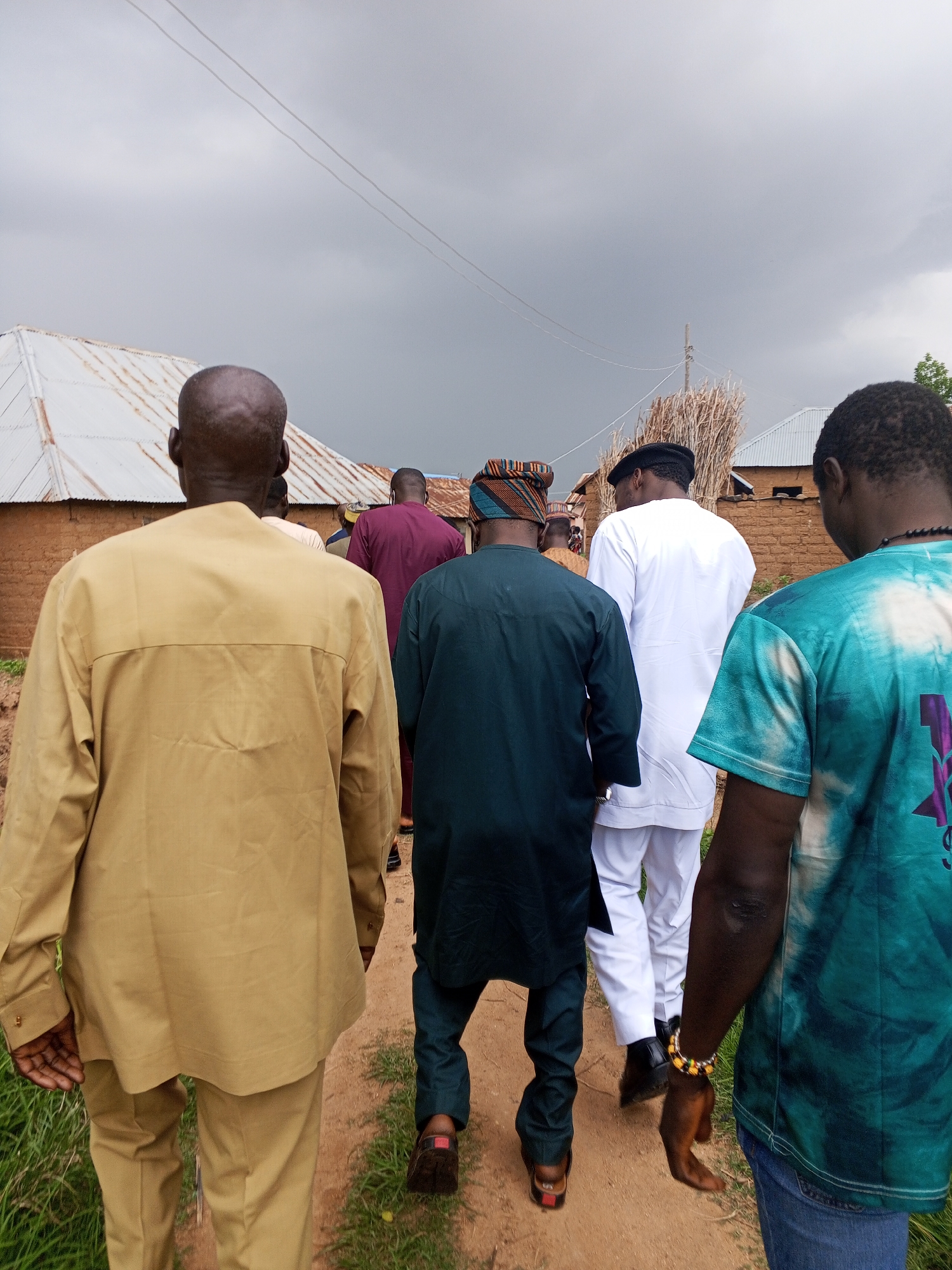 The image captures a group of five men walking away from the camera down a dirt path, which appears to be in a rural or semi-rural area within Panyam, Nigeria. The sky is overcast and grey, suggesting it may be a cloudy day or that rain is imminent.
In the foreground, on the left, a man wearing a tan, long-sleeved outfit and matching pants is walking. His head is shaved, and he is facing away from the camera. Next to him, slightly behind and to the right, is a man in a dark green tunic and pants, also facing away. He has a traditional patterned cap on his head.
Further ahead and to the right of the man in green is a man in a white traditional outfit consisting of a long-sleeved tunic and loose pants. He is also walking away from the camera and has a black cap on his head.
To the far right of the image, closest to the camera, is a man wearing a turquoise and white tie-dye t-shirt and blue jeans. He has a beaded bracelet on his right wrist. His face is not clearly visible, but he is also walking in the same direction as the others.
In the background, there are structures that appear to be houses or shelters, with corrugated metal roofs and mud-brick walls. There are also piles of dry grass or straw visible. The path is bordered by green vegetation on the left side of the image. Power lines are visible stretching across the sky in the upper portion of the image. The overall impression is one of a community gathering or procession taking place in a simple, likely agrarian setting.