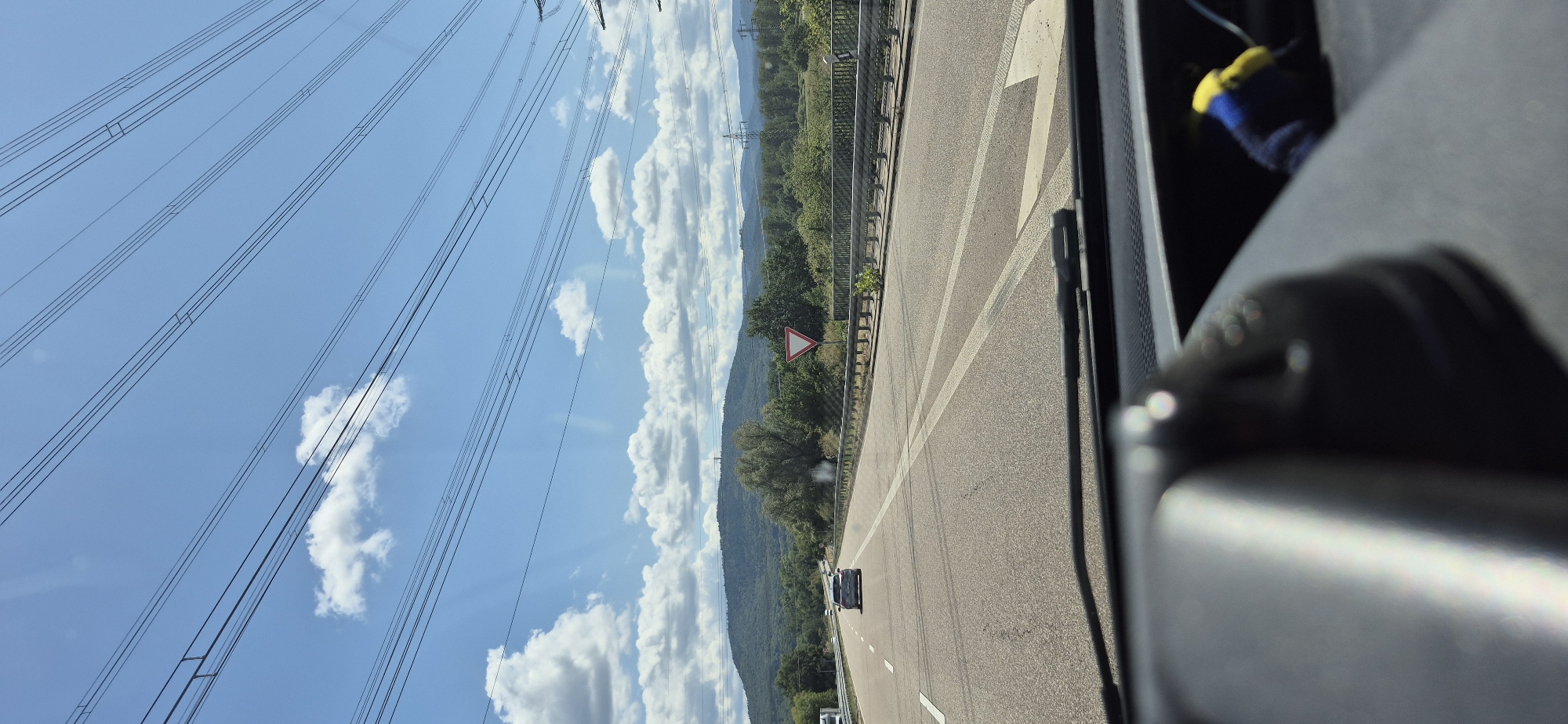 The image shows a view from inside a vehicle driving on a highway in Germany, heading towards a hilly landscape. The sky is blue with scattered white clouds. Multiple high-voltage power lines are strung across the sky. The highway has multiple lanes, with vehicles in the distance. On the right side of the highway, there is a metal guardrail and a triangular yield sign. The landscape in the background consists of a densely forested hill with some buildings or structures visible on its peak. The foreground shows a portion of the car's interior, including the dashboard and part of the steering wheel. The scene appears to be a daytime shot, likely during sunny weather. This location could be on the Autobahn A5, near Baden-Baden, Germany, considering the typical landscape and infrastructure visible in the image.