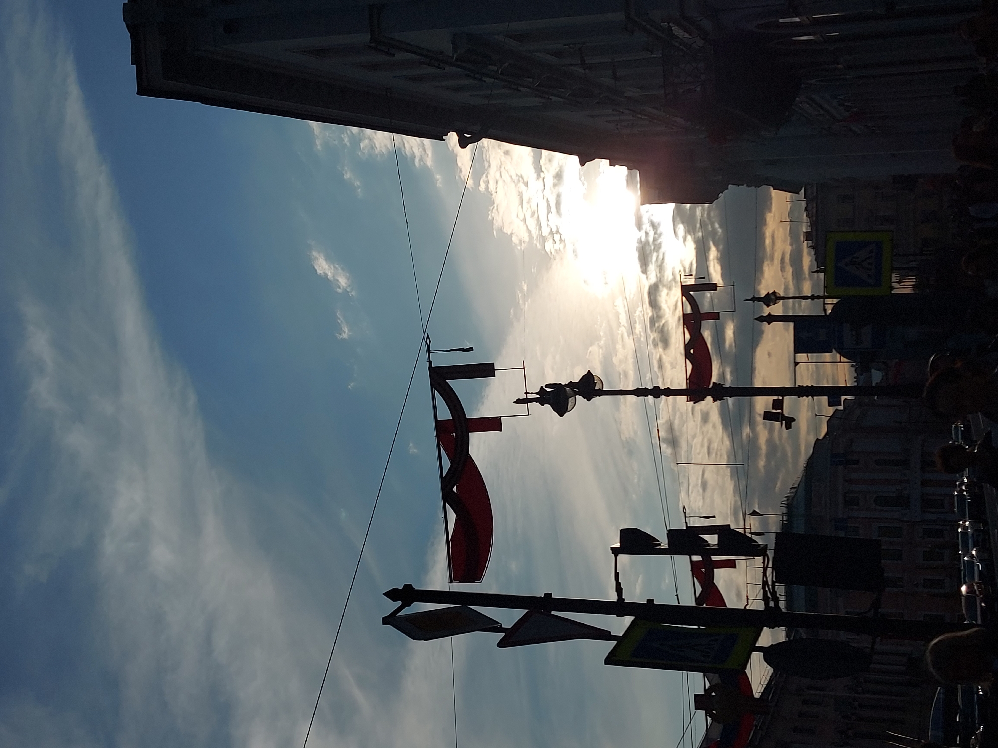 The image shows a street view in Saint Petersburg, Russia, during a sunny day. The scene is captured looking up, with the sky taking up a significant portion. Light clouds fill the blue sky. Silhouetted buildings frame the top of the image. Lamp posts with red flags are visible along the street. There are also street signs, including one for a pedestrian crossing. Silhouetted figures and parked cars are present on the right side of the frame.