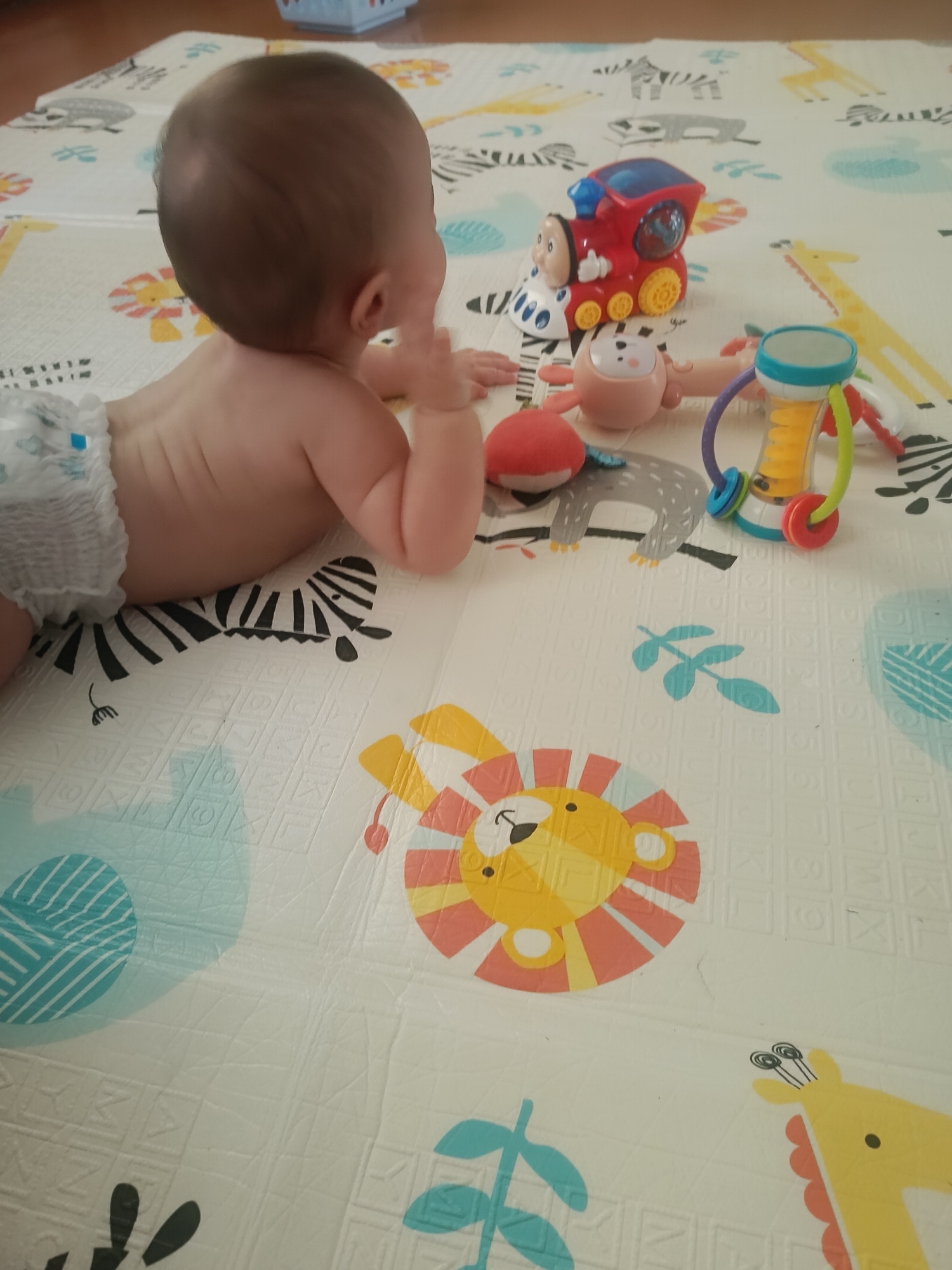 The image shows a baby lying on its stomach on a play mat with cartoon animals. The baby is wearing a diaper and is positioned with its back to the viewer. In front of the baby are several toys, including a red train, a stuffed toy with ears, and a colorful rattle. The play mat has a pattern of various animals, including a zebra, a lion, and a giraffe, as well as some letters of the alphabet. The environment appears to be indoors, possibly a nursery or playroom. The lighting is soft and natural. There is no information in the image to suggest a location in the city of Хохлы, Russia.