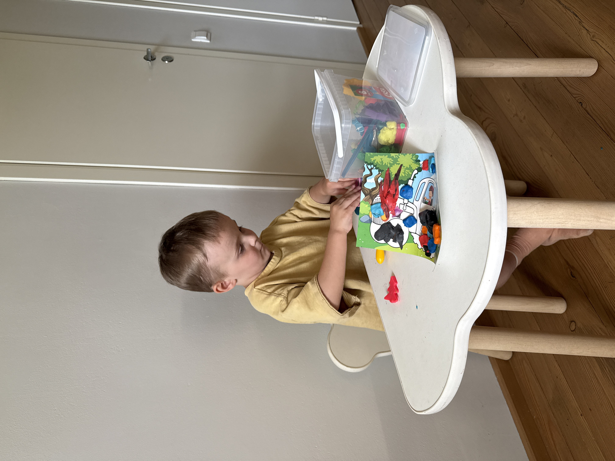 A young Caucasian boy, appearing to be around 3-4 years old, is seated at a white, cloud-shaped table in what seems to be a domestic setting. He is wearing a loose-fitting, mustard-yellow t-shirt. The boy is intently focused on playing with colorful modeling clay. On the table in front of him are several pieces of modeling clay, including red, black, and blue. A green and white children's activity book with images of animals and vehicles is also on the table, along with a clear plastic container filled with various colors of modeling clay. To the right of the container is a transparent plastic lid. The table is supported by three light-colored wooden legs. Behind the boy, a white chair with a curved back is visible. The background shows a light-colored wall and a white door with a silver doorknob and a light switch. The floor is made of polished wooden planks. The scene suggests a child engaged in creative play indoors. The location is Chaikovsky, Russia.