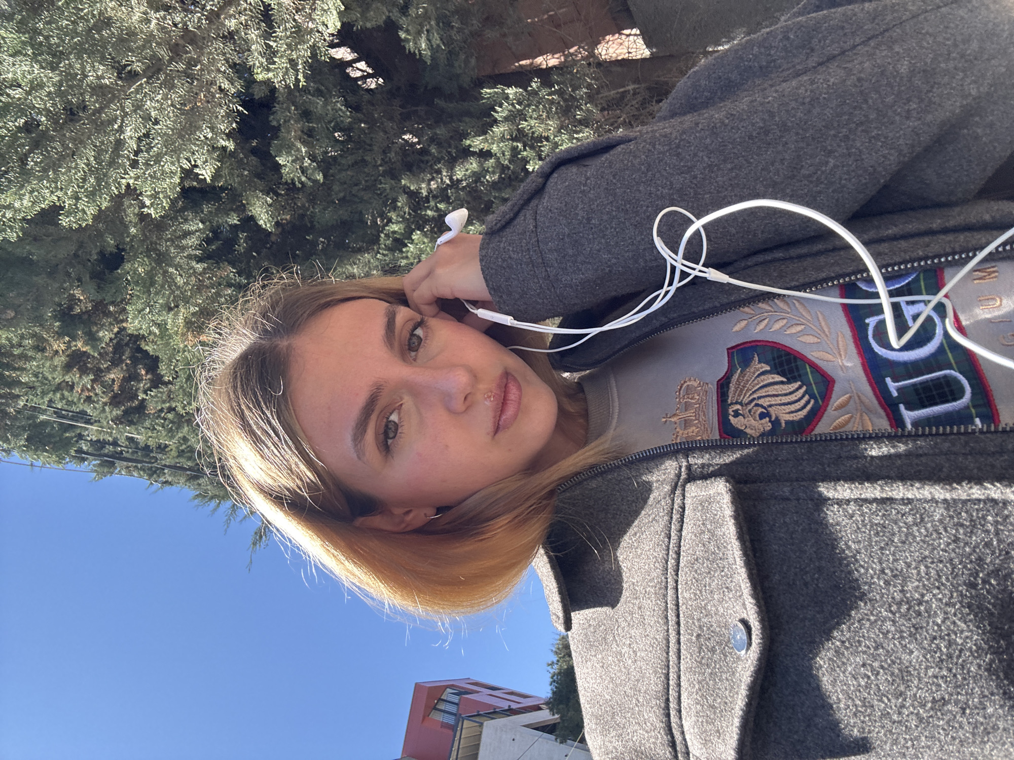 A young woman is outdoors on a sunny day in an urban setting. The sky is clear blue. She is wearing a gray wool coat over a sweater with a collegiate-style design and has white earbuds in her ear. The upper branches of a dense, green tree are in the background. Part of a modern-looking building with red and white sections is visible in the lower left corner of the image.