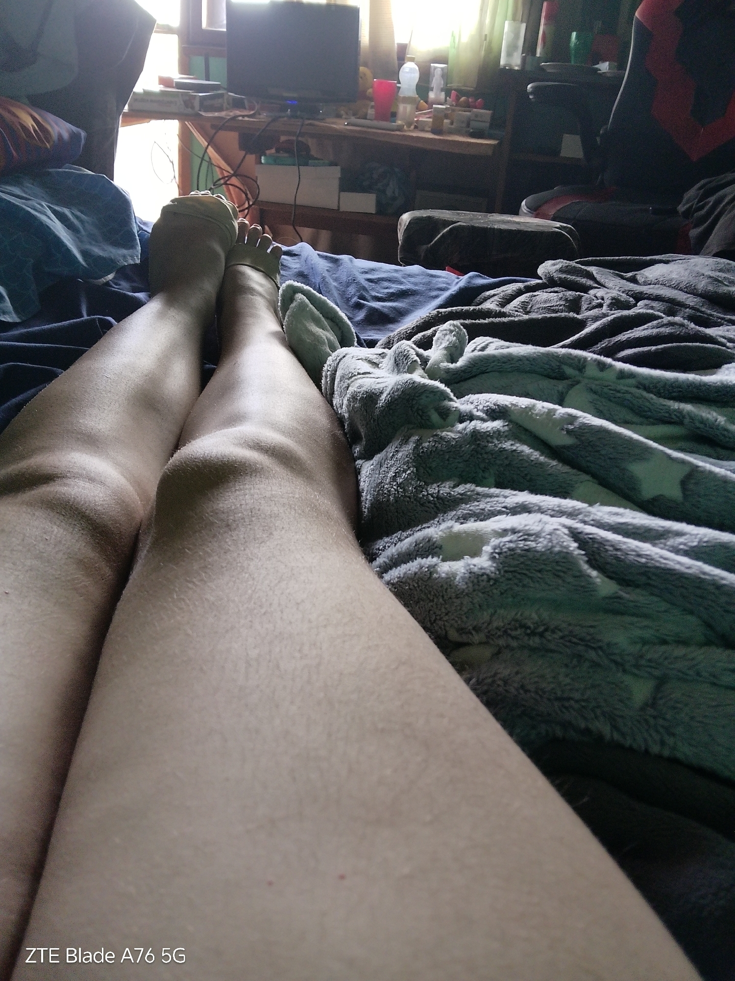 The image shows a close-up view of a person's legs, from the knees down, resting on a bed. The person is wearing beige toe socks. The legs are light-skinned with visible hair. The bed is covered with dark blue sheets and a grey fuzzy blanket with white star patterns. In the background, a desk with various objects, including a television and a gaming chair, is visible. A window with natural light coming through is also present. The scene appears to be an indoor residential setting, possibly a bedroom or living room. No specific location in Stara Zagora, Bulgaria, can be determined from the visual information provided.