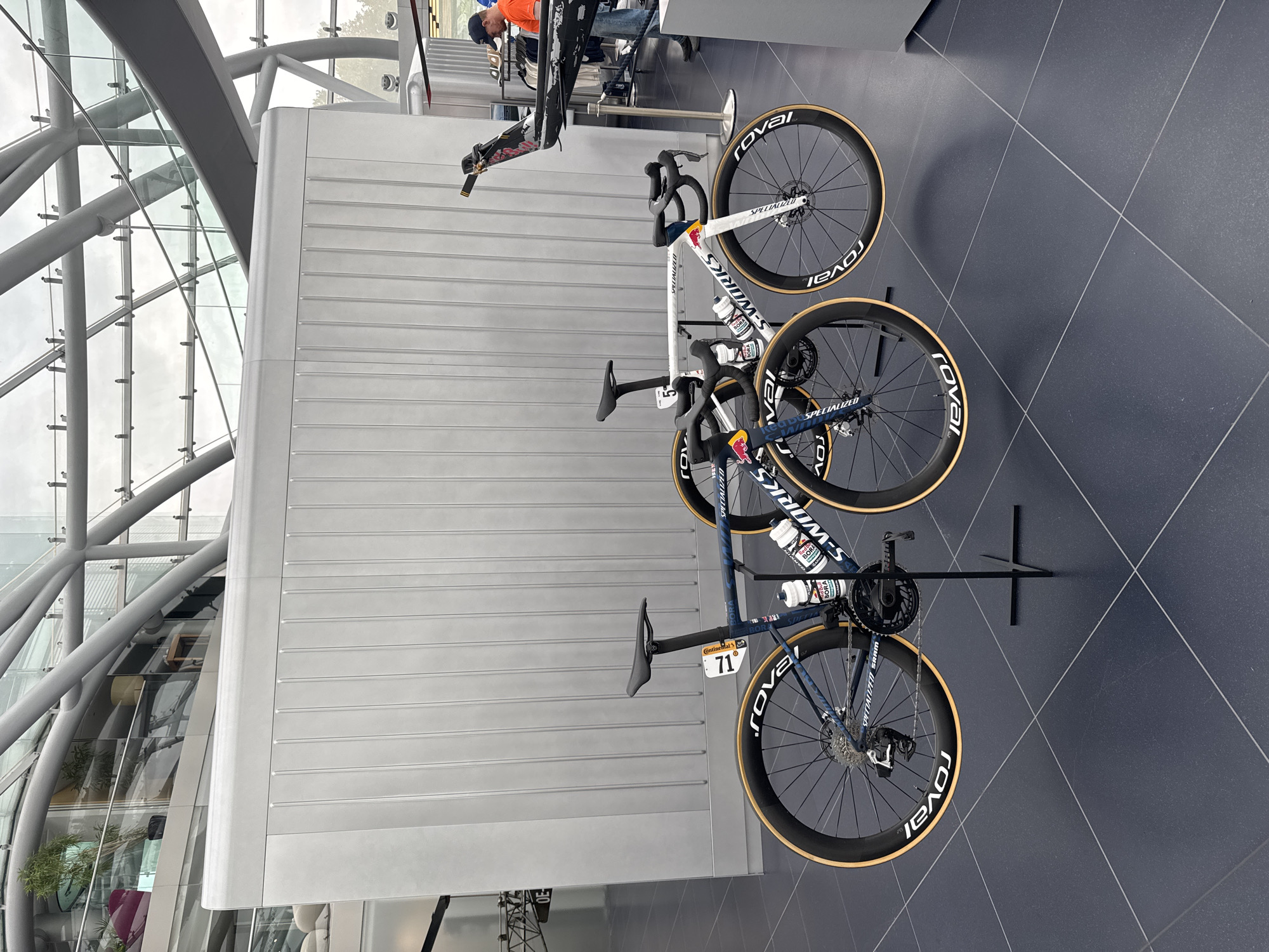 Two Specialized S-Works road bicycles are displayed on black floor stands in a modern indoor space with a metallic, ribbed wall in the background and a glass-paneled ceiling supported by a metal framework. The bicycle on the left is dark blue with a camouflage pattern and a white number "71" on its seat post. It features Roval wheels and a Shimano drivetrain. The bicycle on the right is predominantly white with red and blue accents, and a white number "5" on its seat post. It also has Roval wheels and appears to be a similar model. Both bicycles have water bottles attached to their frames. In the background, a person wearing an orange shirt and a blue cap is partially visible, looking at merchandise on display. The overall setting suggests a showroom or exhibition space, possibly related to cycling or motorsports, given the presence of what appears to be a partially visible aircraft or vehicle component in the far right background. This scene is located within the Hangar-7 at Salzburg Airport, Austria.