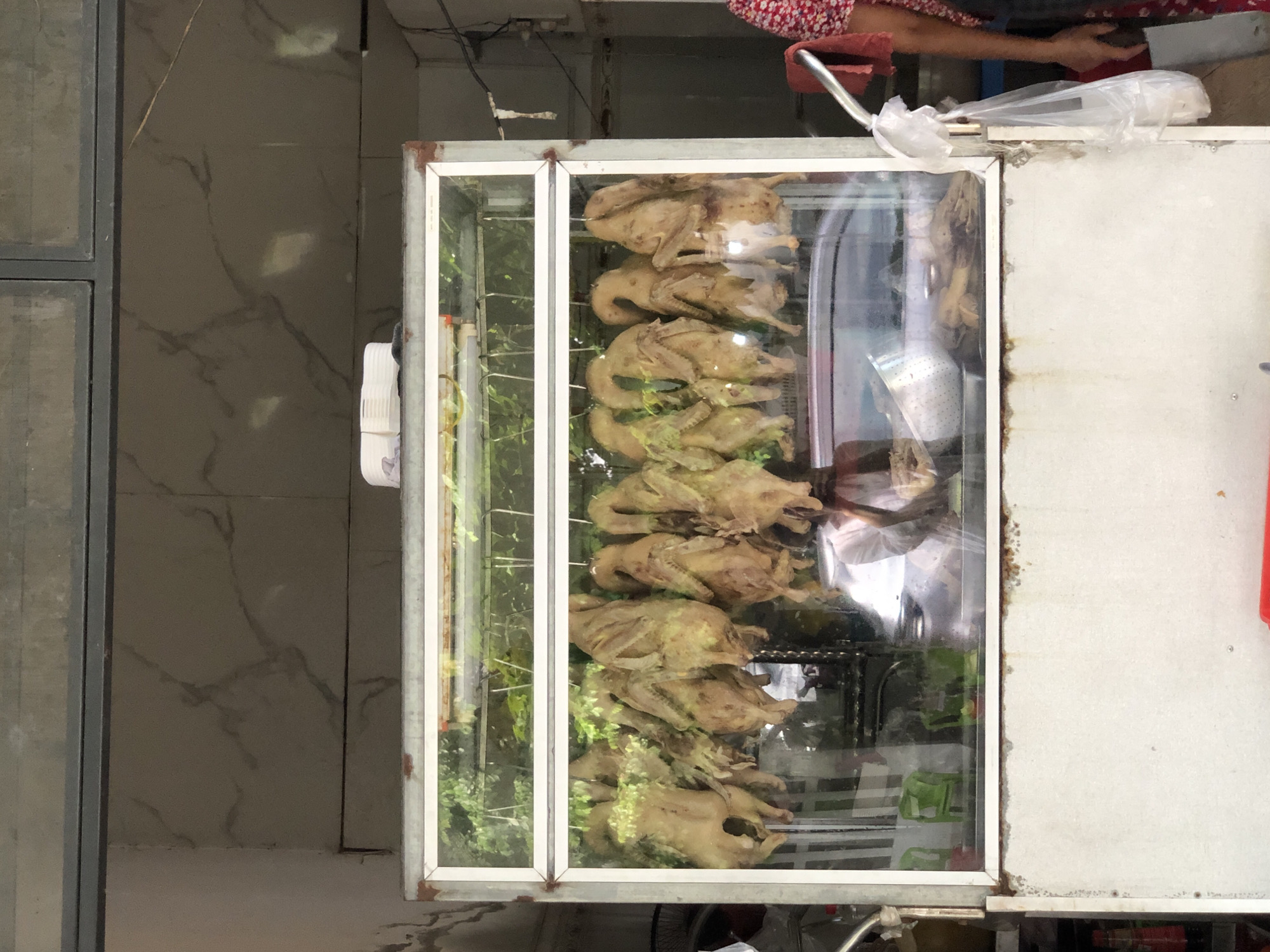 This image depicts a street-side food stall in the city of Đà Nẵng, Vietnam, specializing in roasted duck. A glass-fronted display case is filled with multiple whole roasted ducks hanging from hooks, their golden-brown skin glistening. The ducks appear to be seasoned and possibly glazed, with some visible green herbs stuffed inside their cavities or draped over them. The stall's exterior suggests a busy, outdoor market environment. Behind the display, reflections reveal a street scene, including a parked car and greenery. A person, partially visible on the right side of the image, is attending to the stall, wearing a red and white floral patterned garment. The overall impression is one of a vibrant, authentic local food vendor preparing and showcasing freshly cooked poultry.