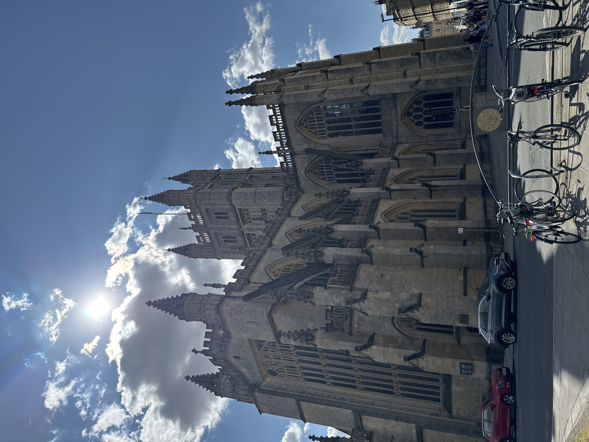 The image shows a low-angle, wide shot of Bath Abbey in Bath, United Kingdom, on a sunny day with scattered clouds. The Abbey, a large Gothic cathedral, dominates the frame with its intricate stone architecture, including spires, arched windows, and flying buttresses. In the foreground, a street runs alongside the Abbey, with parked cars, including a red hatchback and a grey SUV. A bicycle rack with several parked bicycles is also visible on the sidewalk. In the background, other buildings and a few pedestrians are visible. The sun is shining brightly from behind a cloud, creating lens flare and casting shadows.