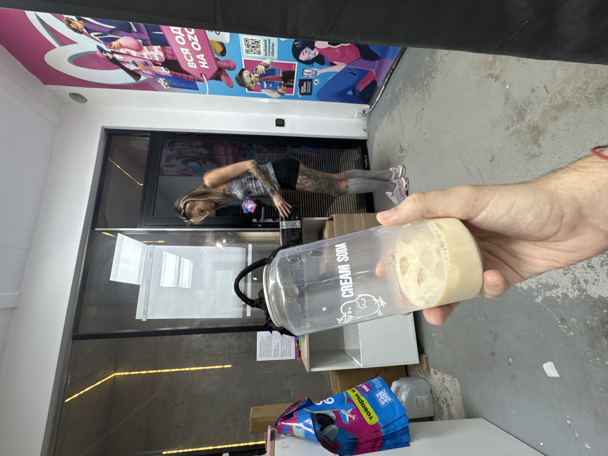 A person's hand holds a clear plastic cup filled with a light brown liquid and ice cubes. The cup has a black braided handle and the words "CREAM SODA" printed in white, along with a cartoon chick graphic. In the background, a woman with tattoos on her arms and legs, wearing a grey tank top, black shorts, and grey socks, stands near a glass door. She appears to be interacting with a black box on a shelf. To the right, a large pink and blue advertisement poster features stylized cartoon characters. The scene takes place indoors, likely in a commercial or retail space. Yellow string lights are visible on the wall behind the woman.