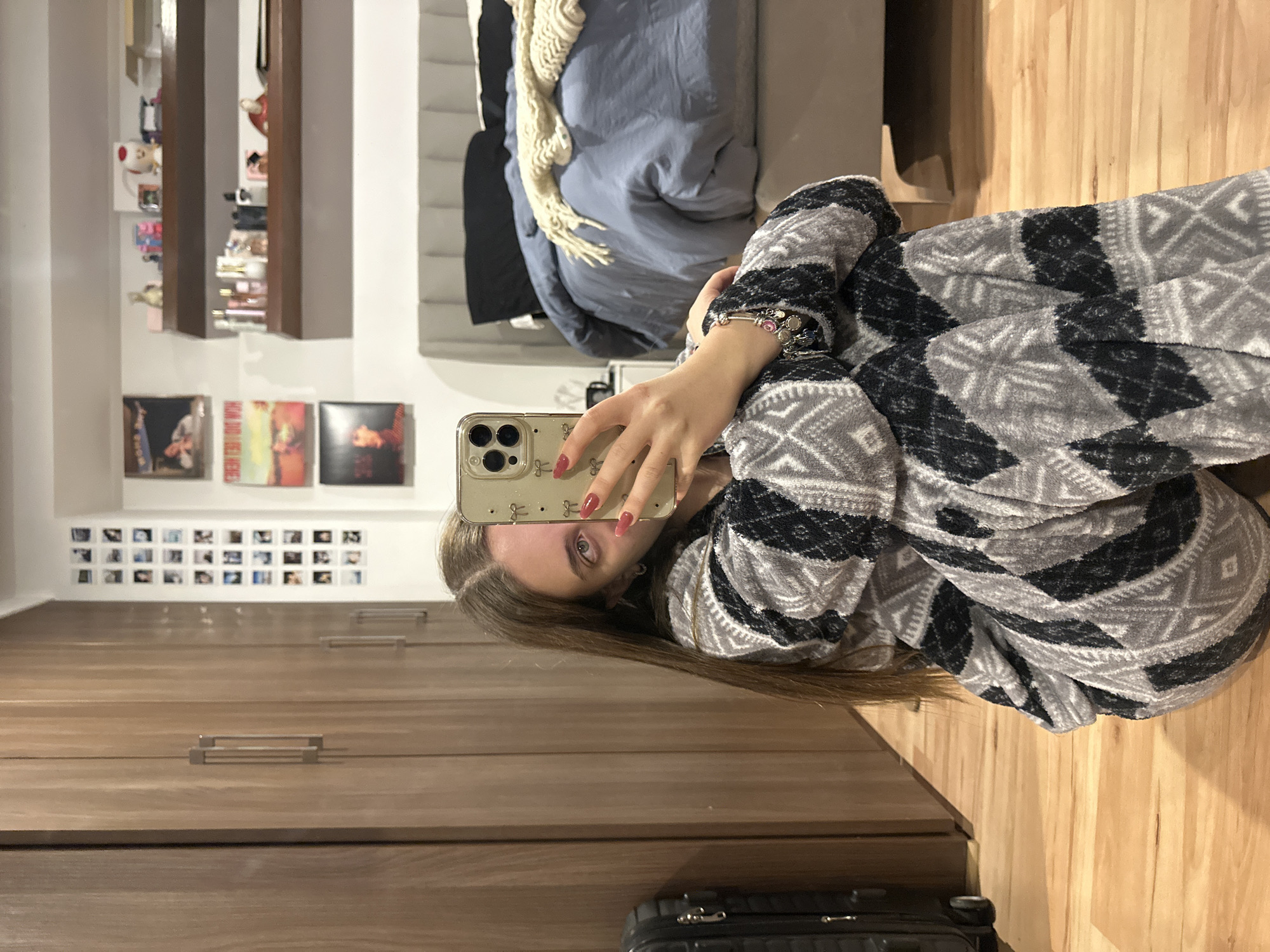 A young woman takes a selfie in a bedroom setting. The woman is wearing a gray and black patterned pajama set and holding a smartphone with a light-colored case. Her fingernails are painted red. The bedroom features a wooden floor and a cabinet or dresser with horizontal wood grain. On the wall above the dresser are small square photographs and a shelf with various items. In the background is a bed with blue bedding and a white throw. A suitcase is on the floor near the dresser. The image does not provide enough information to determine if this location is in Huixquilucan de Degollado, Mexico.