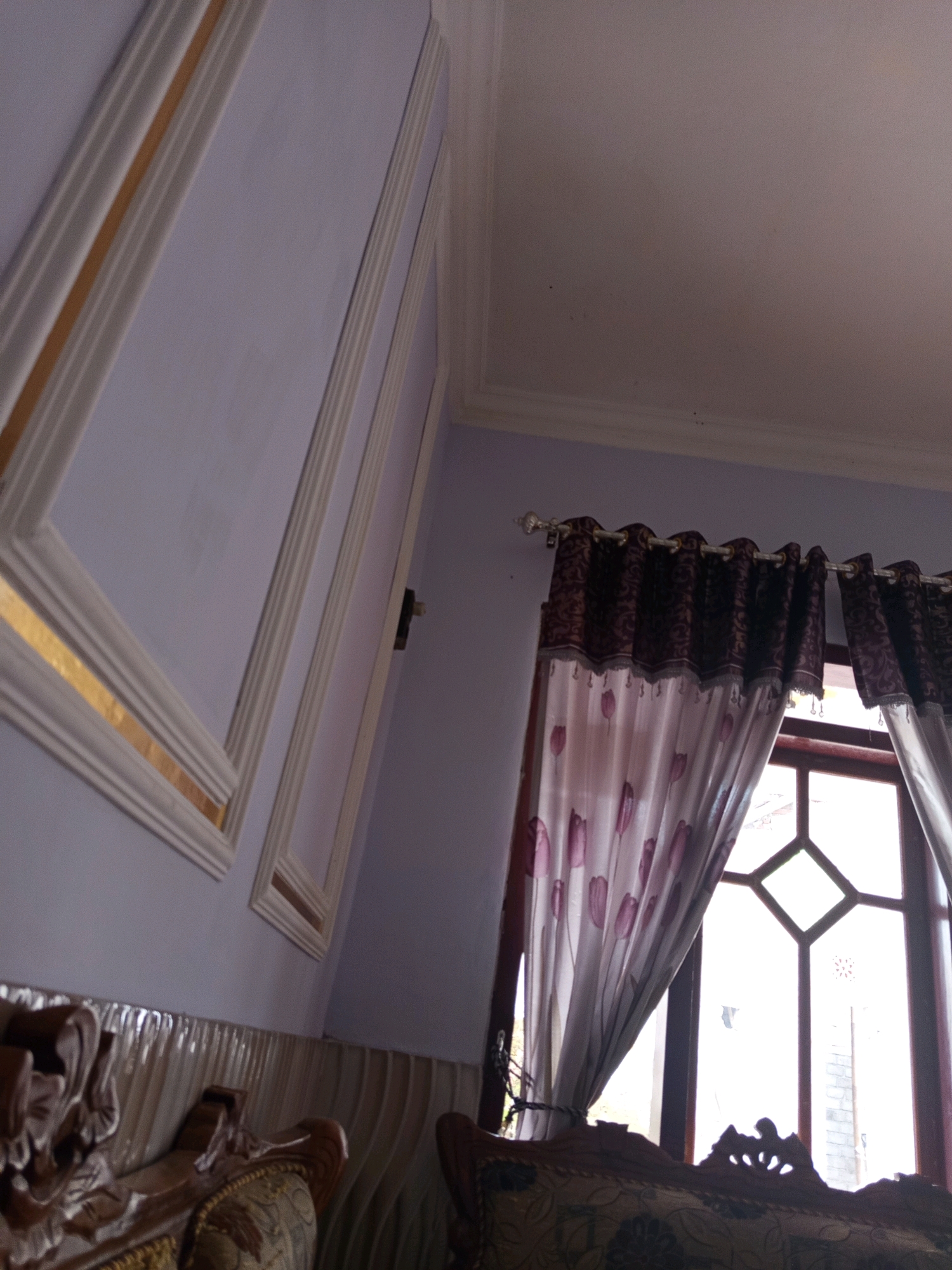 The image captures an interior scene, likely a living room, with a focus on decorative elements and natural light. The left side of the frame features a wall with ornate white trim and a thin gold accent strip, creating a geometric design. The wall itself is painted a soft lavender color. In the background, a large window is partially covered by patterned curtains. The curtains have a dark, intricate floral or damask pattern at the top and a lighter, sheer fabric with a repeating tulip motif towards the bottom. The window frame appears to be made of dark wood. The light streaming through the window suggests it is daytime. In the foreground, the armrest of a dark wooden sofa with carved detailing is visible. The sofa is upholstered in a patterned fabric. The overall impression is one of a well-decorated, possibly traditional interior space. No specific location in Rowo Indah, Indonesia, can be identified from the visual information alone. There are no people or actions depicted.