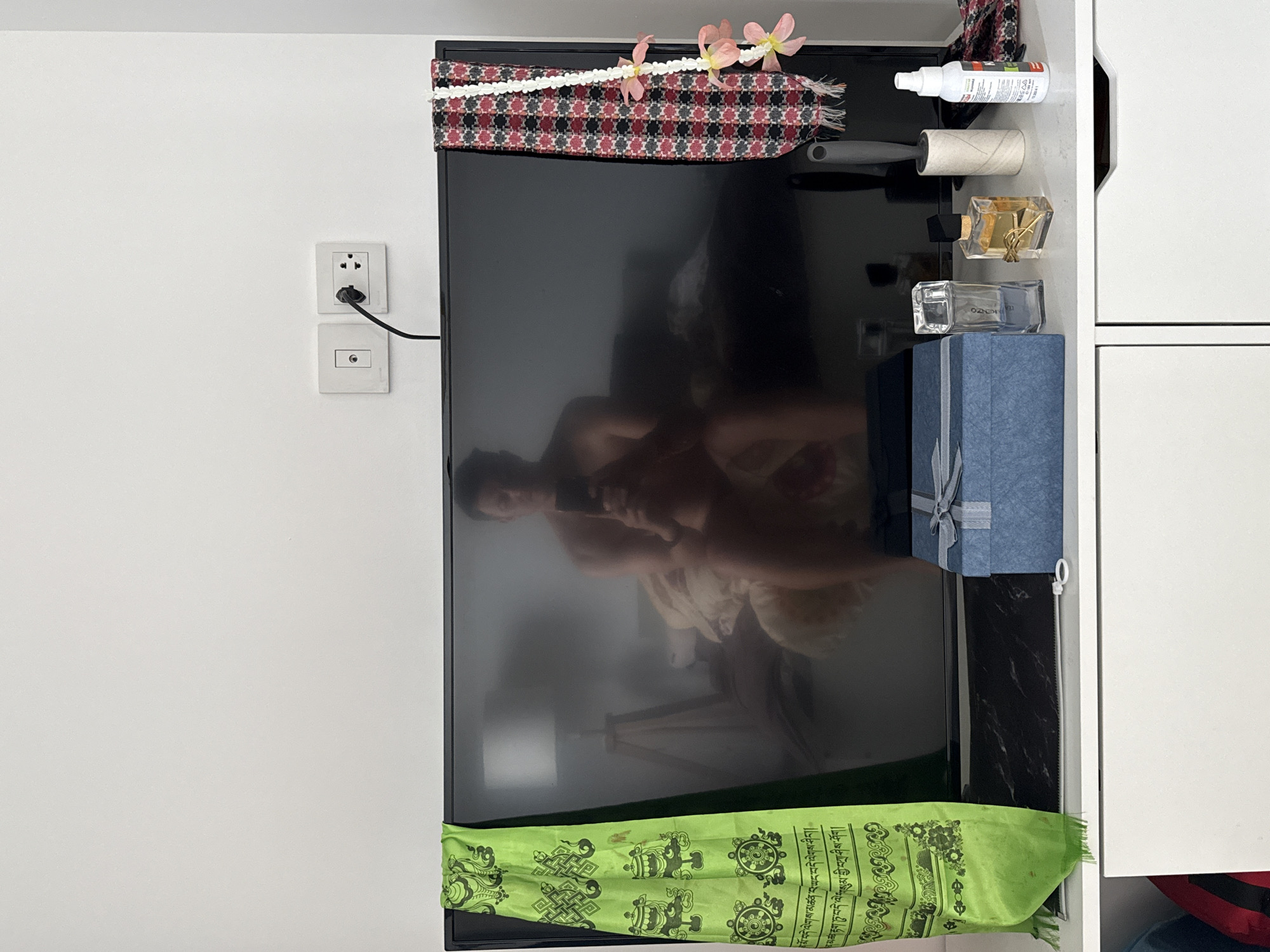A vertical medium shot reveals a living room interior in Bangkok, Thailand, with a television as the central object. The TV screen reflects a blurred image of a person using a mobile device, likely taking the photograph.
On top of the TV is a colorful, patterned piece of fabric, adorned with a string of pink flowers. Adjacent to the TV, on the right side, is a cabinet filled with assorted items: a white spray bottle, a roll of lint remover, two perfume bottles, and a blue gift box with a silver ribbon.
A bright green cloth with intricate designs drapes along the bottom of the TV. To the left of the TV, on the wall, is a double electrical outlet. The overall scene suggests a lived-in, personal space within an urban environment.