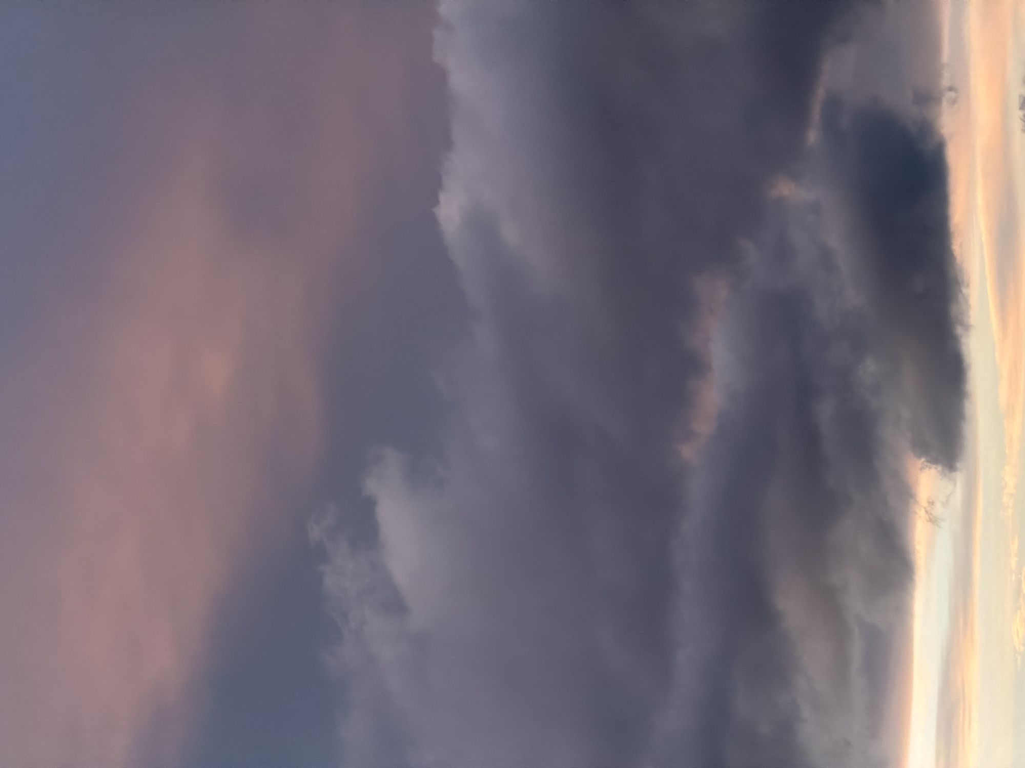 The image presents a view of the sky at dusk or dawn. Dominating the scene are dark, billowing storm clouds. Patches of soft orange and pink light peek through gaps in the clouds, suggesting the position of the sun just below the horizon.
The image is likely from Bragança, Portugal, based on the prompt's context. No people or man-made objects are visible. The environment consists of the sky and clouds with the sun providing a backdrop. The interaction consists of the interplay between the dark clouds and the light of the sun.