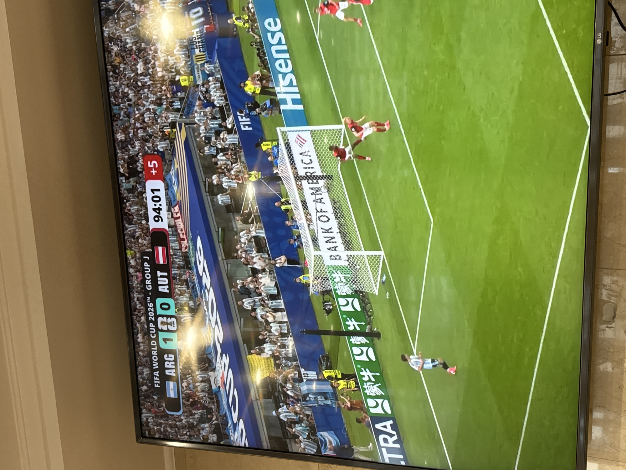 This image depicts a television screen displaying a soccer match. The score shows Argentina (ARG) leading Austria (AUT) 1-0. The match is in the 94th minute with 5 minutes of added time. The game is identified as FIFA World Cup 2026™, Group J. The screen shows the green turf of a soccer field, with players in red and white jerseys in action near a goal. Spectators are visible in the background, wearing white and light blue attire, suggesting support for Argentina. Commercial advertisements for "BANK OF AMERICA" and "Hisense" are displayed on the screen. The television is set on a tiled surface, presumably in a residential setting in North Bergen, United States of America, given the context of the provided location.