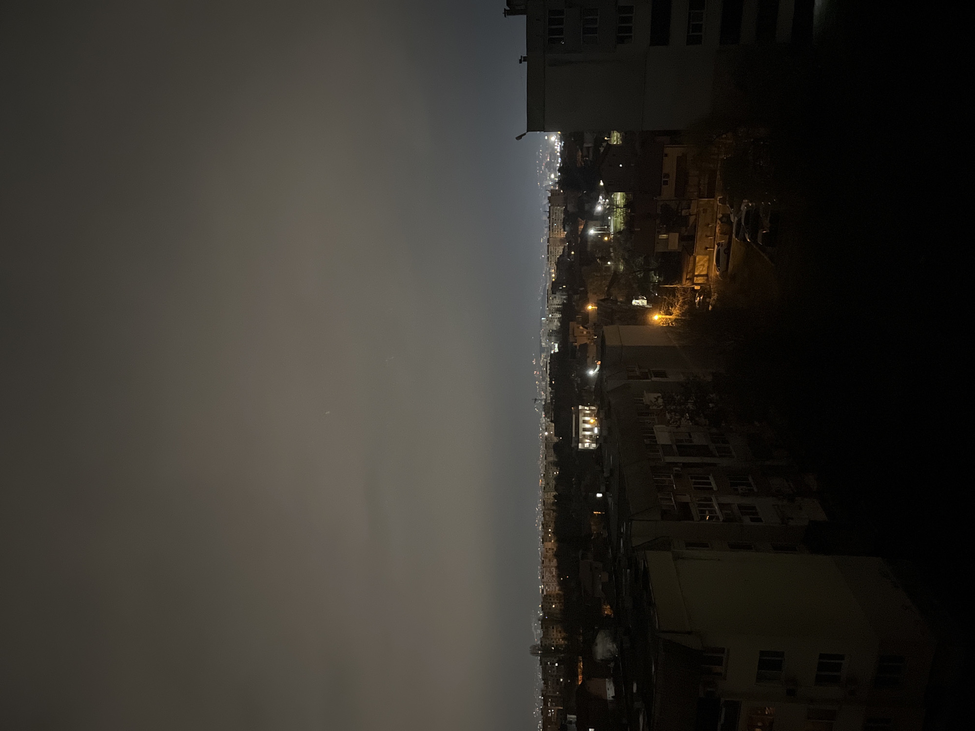 The image presents a nighttime urban scene, likely in Sofia, Bulgaria. It's a high-angle, horizontal shot capturing a city skyline partially obscured by a layer of smog or fog.
**Environment:** The left half of the image is dominated by a dark, overcast sky, appearing dense with particulate matter or atmospheric haze. The right half shows a lit-up city area, though the lights are somewhat diffused due to the atmospheric conditions. Building silhouettes and streetlights are visible, and the overall illumination appears subdued, contributing to a hazy, low-visibility environment.
**Objects:** Buildings of varying heights are present in the city portion of the scene. Streetlights illuminate the streets.
**People/Subjects/Actions/Interactions:** Not discernible in this image due to the angle, atmospheric conditions, and time of day.