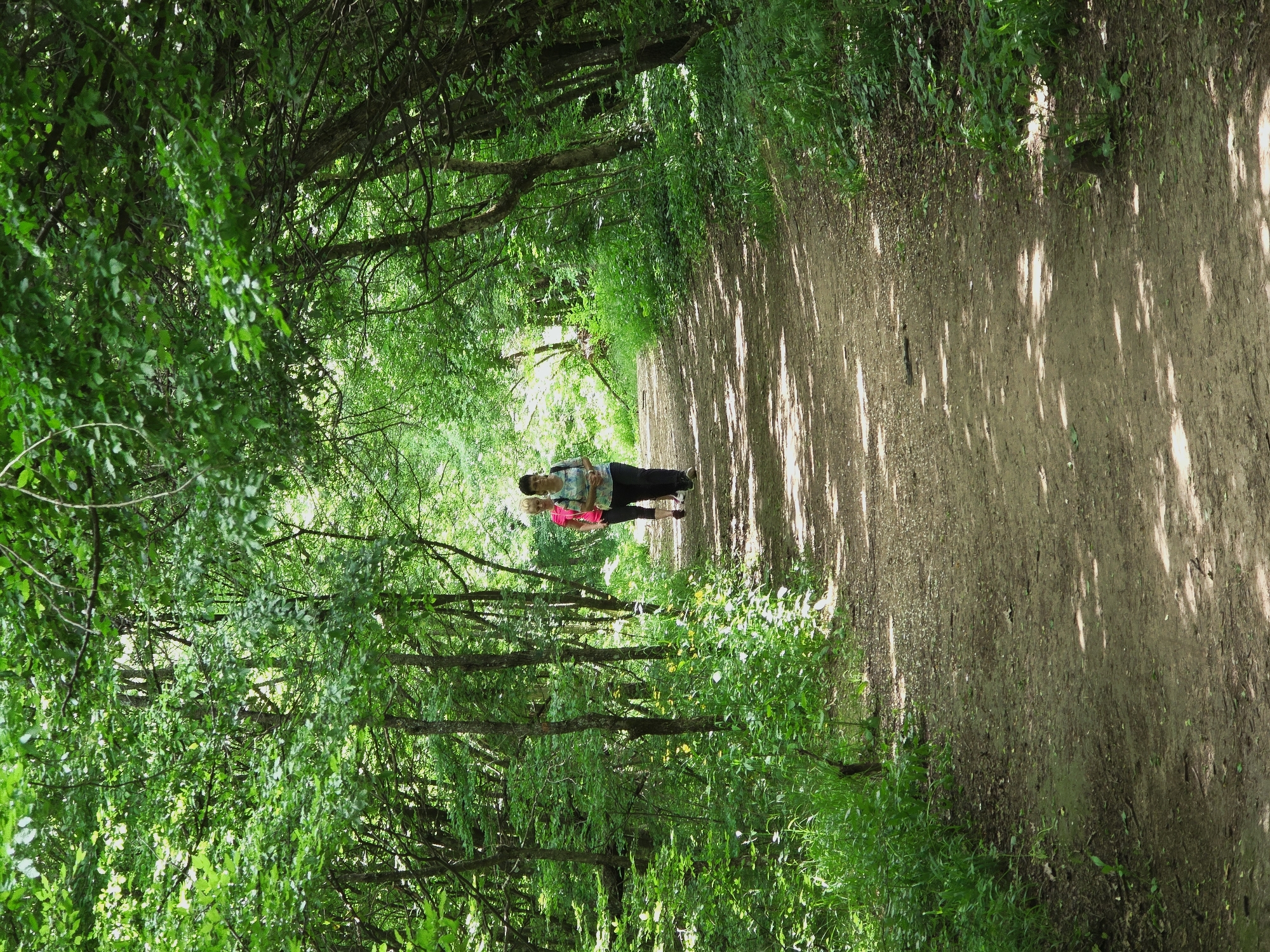 Two people stand on a dirt path lined with trees in a forest. The trees create a dense canopy overhead, casting shadows on the path. The path is earthen and appears well-worn. The location is the city of Shumen, Bulgaria. One person wears a pink top, and the other wears a blue top. They are standing close together.
