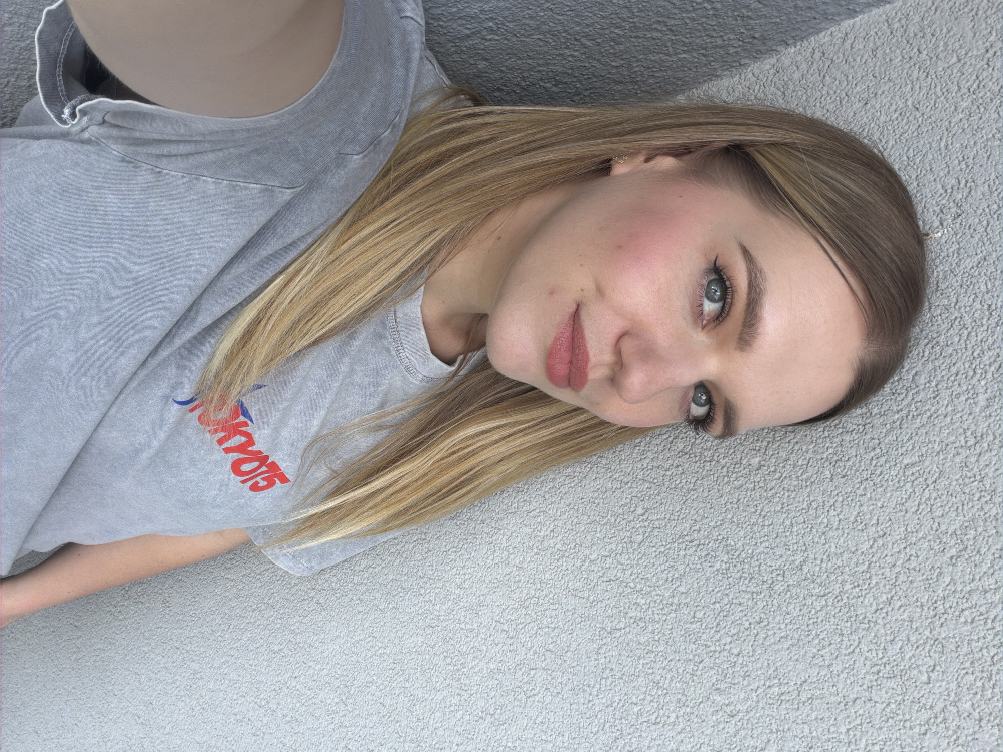 A woman with blonde hair, blue eyes, and fair skin poses for a selfie. She is wearing a grey t-shirt with "TOKYO15" printed on it in red and blue lettering. She has on makeup including eyeliner, blush, and lipstick. She is standing in front of a textured grey wall. The lighting suggests it is daytime. The image does not provide enough information to determine a specific location within Riga, Latvia.