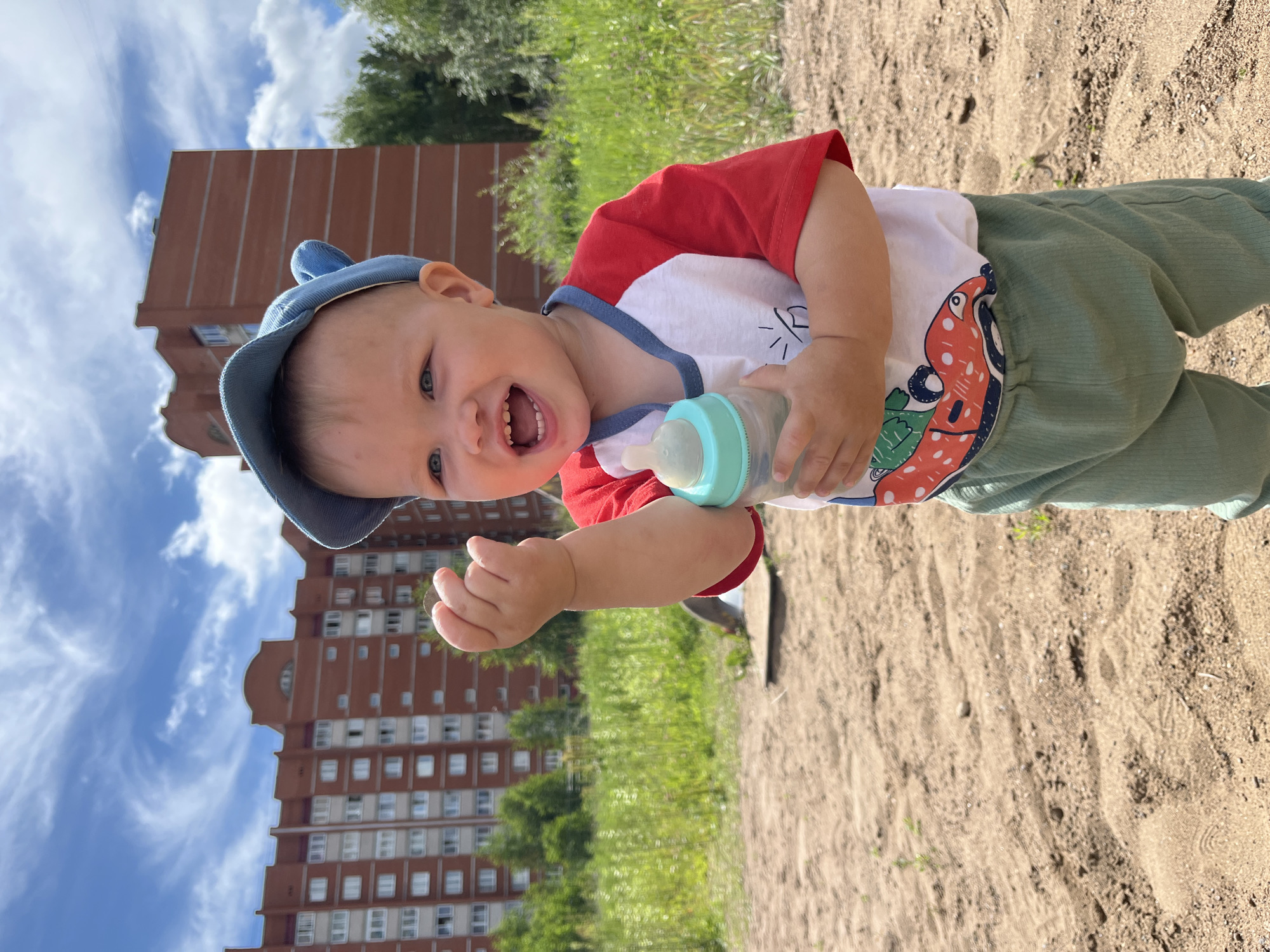 In the city of Chaikovsky, Russia, on a bright, cloudy day, a young toddler stands on a sandy surface, likely a playground. The toddler, wearing a blue cap with bear ears, a red and white raglan shirt with a graphic print, and light green pants, is holding a baby bottle with a turquoise cap. The toddler's mouth is open in a wide smile, and their left hand is raised. In the background, a tall, red brick apartment building stands tall against the blue sky with wispy white clouds. Green grass and trees are visible on either side of the building, suggesting an urban setting with accessible green spaces. The sand in the foreground shows evidence of footprints and small patches of dry grass, indicating a well-used outdoor area.