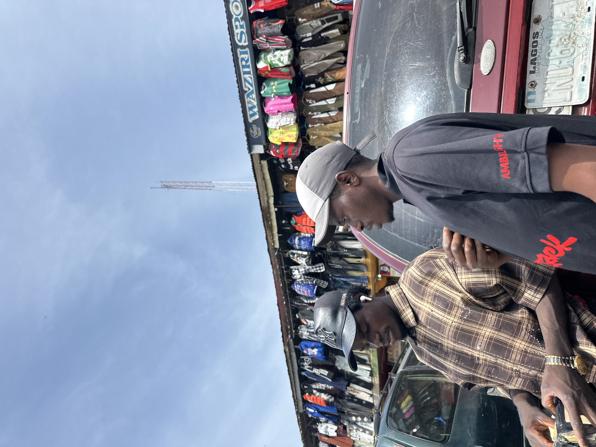 This media file captures two men in an outdoor market setting in Jos, Nigeria. The men are the primary subjects, both wearing baseball caps and casual attire. One man, facing the camera, is wearing a dark t-shirt with red lettering that reads "AMBILIGHT" on the sleeve, and a grey baseball cap. The other man, in the foreground and facing away, wears a brown plaid shirt and a black baseball cap with "NEW YORK" embroidered on it. He is also wearing a gold watch.
The background reveals a stall selling clothing, with numerous items like jerseys and jackets hanging on display. The sign above the stall reads "WAZIRI SPORT". To the right, a maroon vehicle is visible, featuring a license plate that indicates "LAGOS, FEDERAL REPUBLIC OF NIGERIA". The scene appears to be taking place during the day, under a bright, overcast sky. The overall atmosphere suggests a typical market day, with people engaging in commerce. The orientation of the image is rotated 90 degrees clockwise.