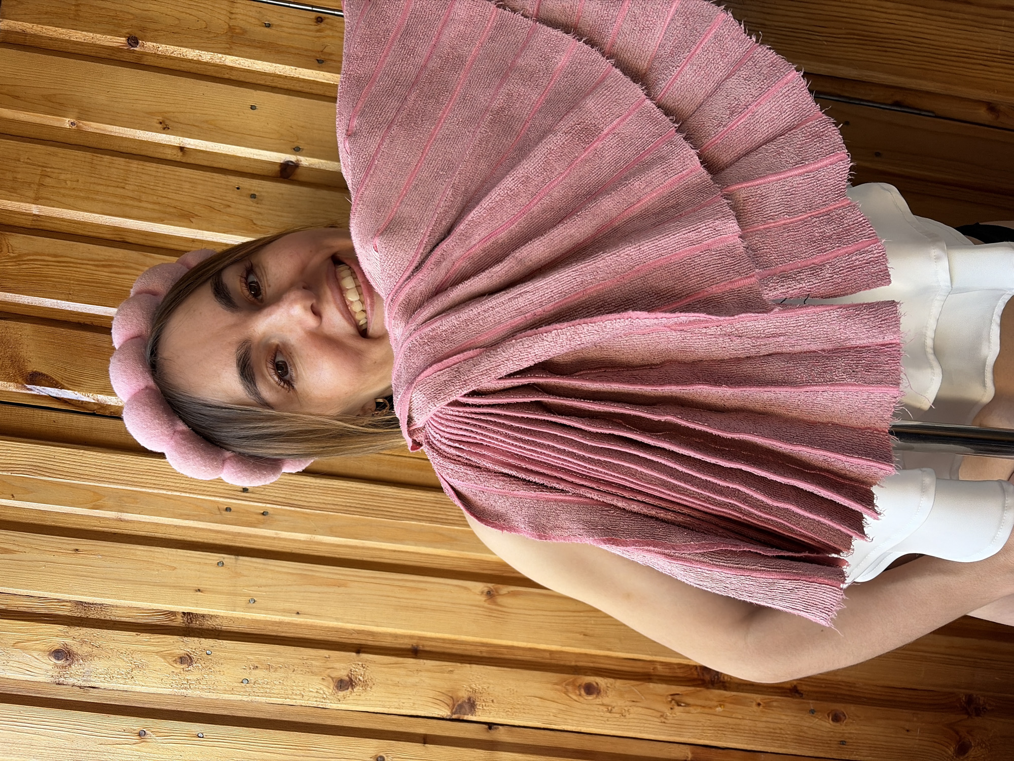 A woman is shown from a low-angle perspective against a backdrop of horizontal wooden planks, possibly within a sauna or wood-paneled room. The planks vary in color from light to medium brown, displaying natural wood grain and knots. The woman has fair skin, brown hair, and is smiling, revealing her teeth. She is wearing a pink, padded headband that matches the color of a folded, textured pink cloth or towel she holds in front of her face. The cloth is arranged in overlapping layers, creating a fan-like effect. A portion of her upper body is visible, including a white garment with a visible collar. She is also wearing what appear to be white wrist supports. The subject is holding onto a chrome pole.