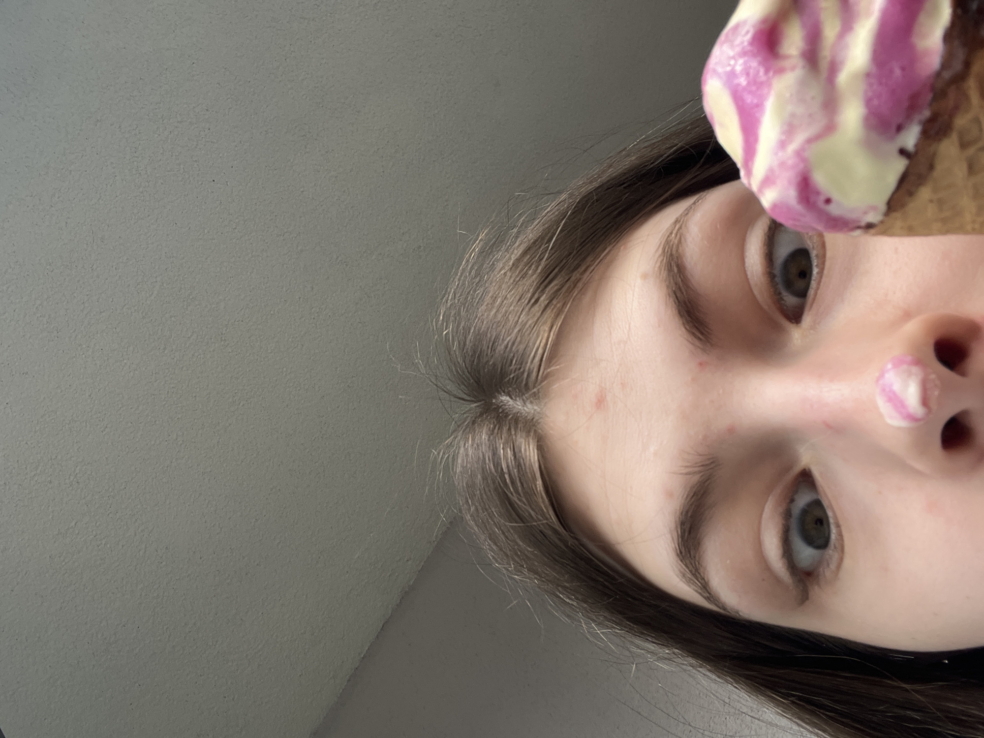 An extreme close-up, low-angle photograph captures a young individual indoors in Saint Petersburg, Russia. The individual, with long, dark brown hair parted centrally, is seen from the forehead down to the upper chest. Their wide-open eyes gaze upwards towards the camera, with the pupils slightly dilated. The fair skin exhibits minor reddish blemishes on the forehead. A small, irregular glob of creamy white and magenta-swirled substance, identified as ice cream, rests on the tip of the nose. In the lower right foreground, a waffle cone holds more of the same two-toned, swirled ice cream. The background is a plain, light grey, textured ceiling or wall, with a discernable corner where surfaces meet in the upper left. The environment is illuminated by even, diffuse lighting.