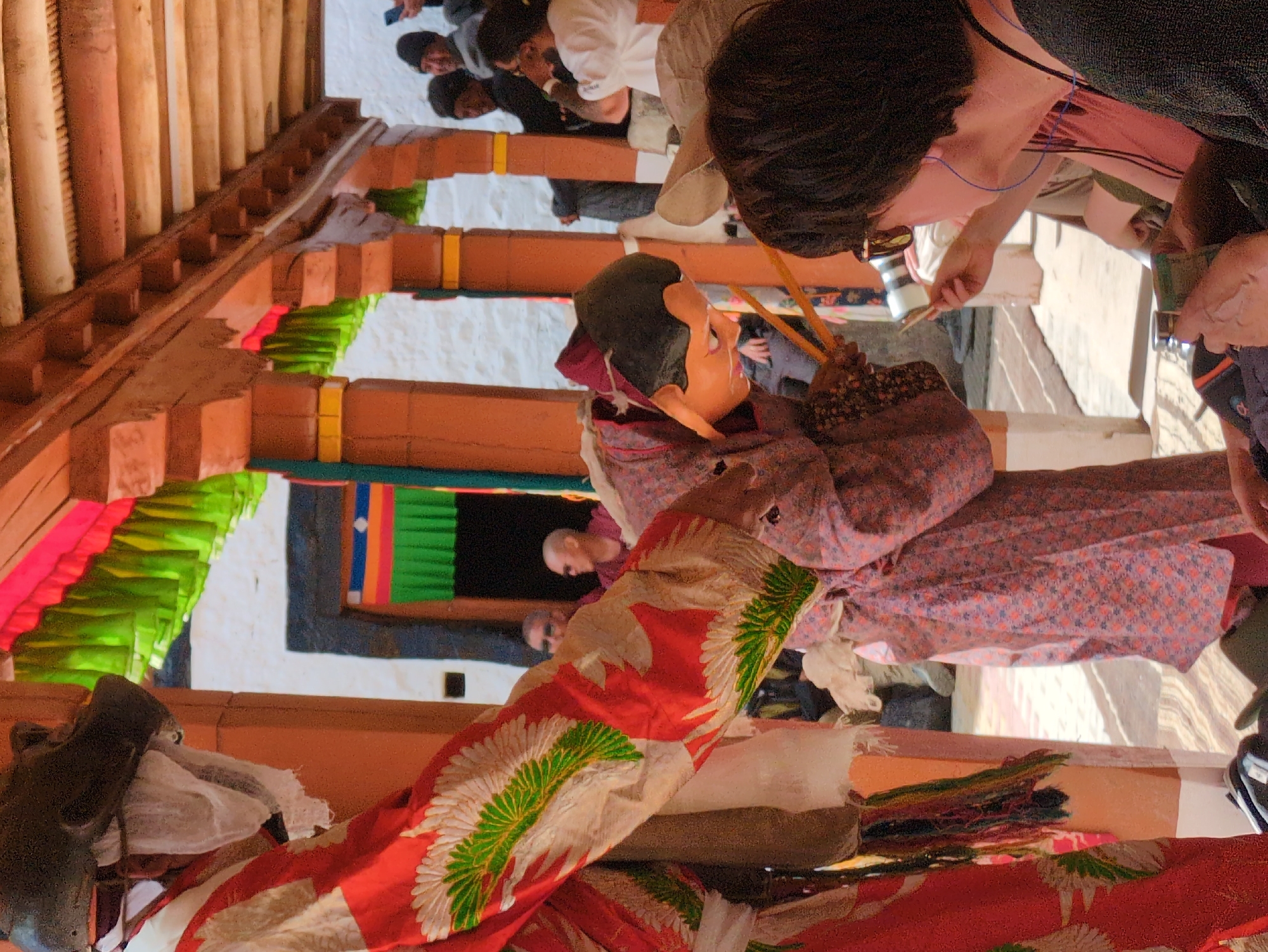 In Pibiting, India, a traditional masked dance performance takes place under a wooden canopy adorned with colorful fabric streamers. Two individuals in elaborate costumes are the central figures. One, on the left, wears a large, dark, animal-like mask and a vibrant red robe decorated with white and green feather-like patterns. The other, on the right, is partially visible, wearing a patterned pink robe and a tan mask with dark hair, holding two thin wooden sticks. They appear to be engaged in a ritualistic dance, with the masked figure on the right seemingly in motion. A crowd of onlookers is gathered behind them, observing the performance. Among the spectators, a person in the foreground is wearing sunglasses and holding a small object. The environment suggests an outdoor courtyard or temple setting, with white walls and wooden pillars supporting the roof structure. The ground is paved with a textured surface.