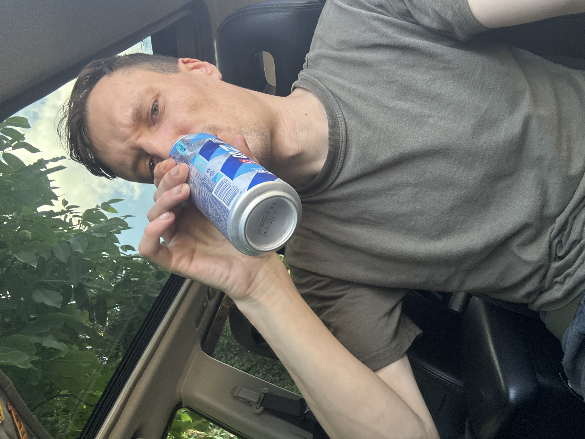A person, identified as male, is seated inside a vehicle, holding a beverage can to his lips and drinking. He is wearing a plain, olive-green t-shirt. The interior of the vehicle is visible, including a black leather seat and the sunroof, through which green foliage and a light blue sky are seen. The scene is lit by natural daylight. The specific location within the city of Shatava, Ukraine, is not discernible from the image.