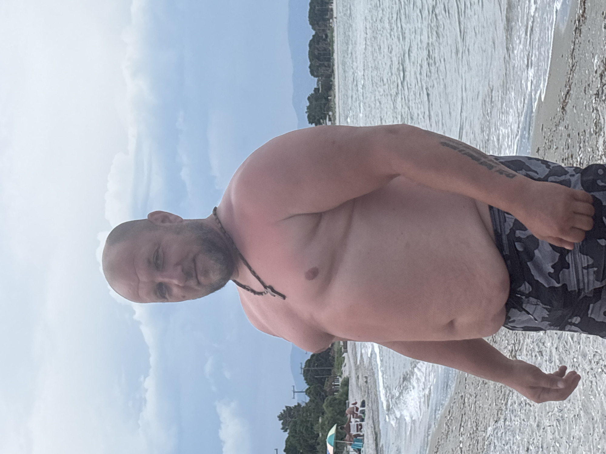 A middle-aged, Caucasian man with a shaved head and beard stands on a beach, facing the camera. He is wearing camouflage swim trunks and a necklace with a cross pendant. The man appears to be sunburnt on his upper body. The background shows the Aegean Sea with gentle waves, a pebbly shore, and a line of trees and distant hills. There are some beach umbrellas and people in the distance to the left of the frame, suggesting a public beach. The sky is overcast. The image was taken in Mandra, Greece.