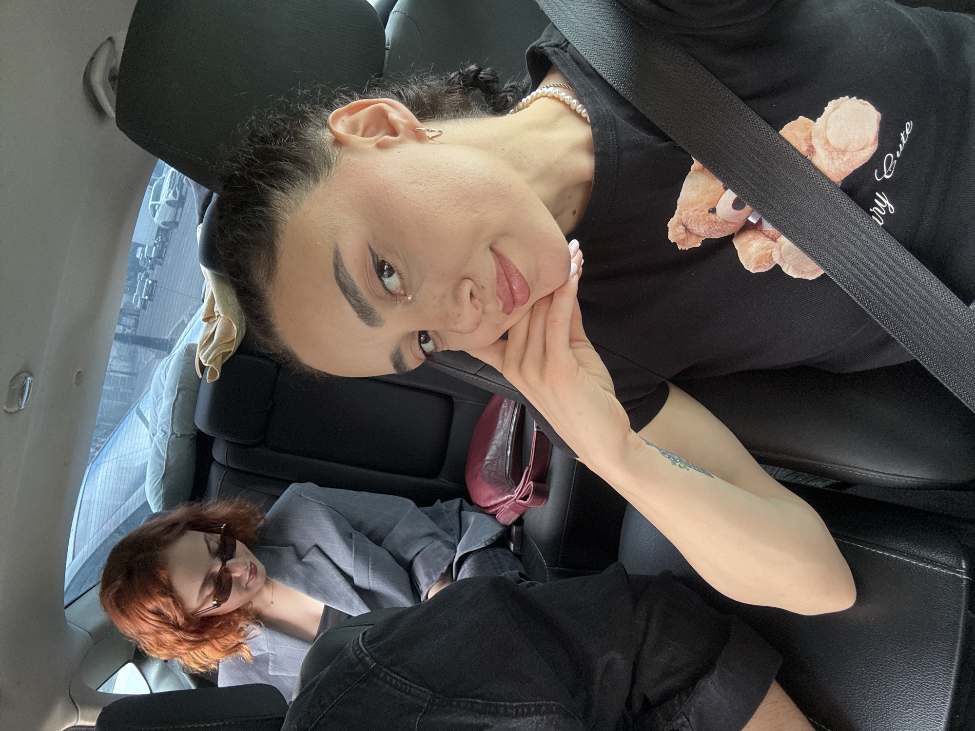Two women are in the back seat of a car. The woman in the foreground is sitting up and facing the camera. She has dark hair pulled back, fair skin, and is wearing a black shirt with a teddy bear design, and a pearl necklace. A seatbelt crosses her chest. Her left hand is propped under her chin. The second woman, who appears to have red hair, is lying down with her head near the car window. She is wearing a gray blazer and sunglasses. A burgundy purse is positioned between them on the seat. The background suggests the scene is in a city, possibly Tbilisi, Georgia, based on the urban setting visible through the car window.