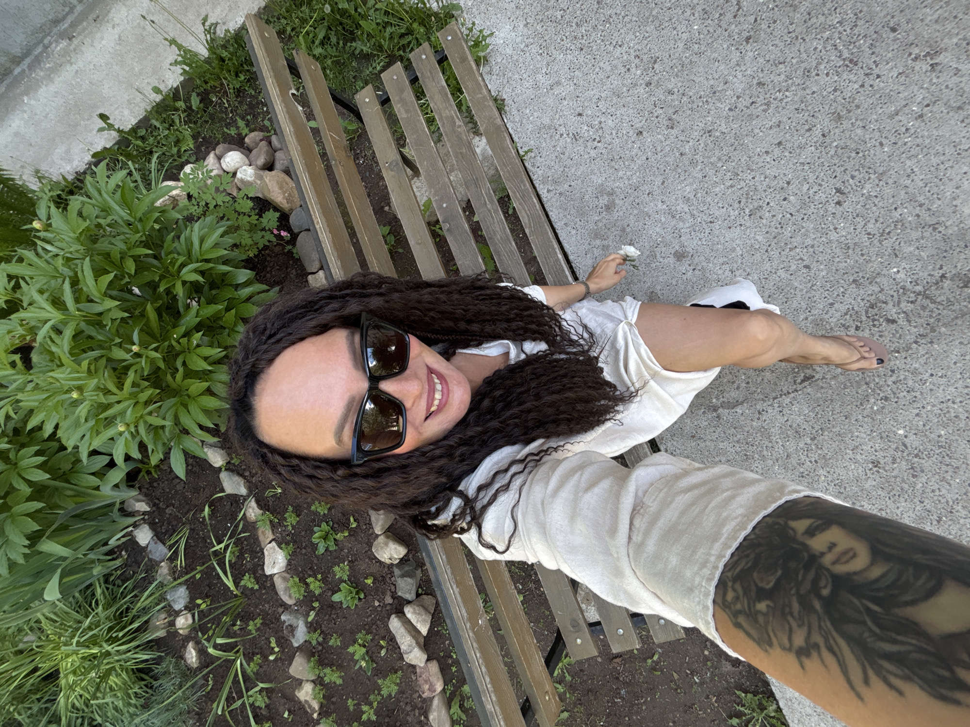 A woman with long, dark, curly hair and wearing sunglasses is sitting on a wooden bench next to a garden. The woman is fair-skinned and is wearing a light-colored, possibly white, outfit. A tattoo is visible on her arm. The garden features green plants and small rocks. A concrete walkway is next to the bench. The location is in the city of Chaikovsky, Russia.