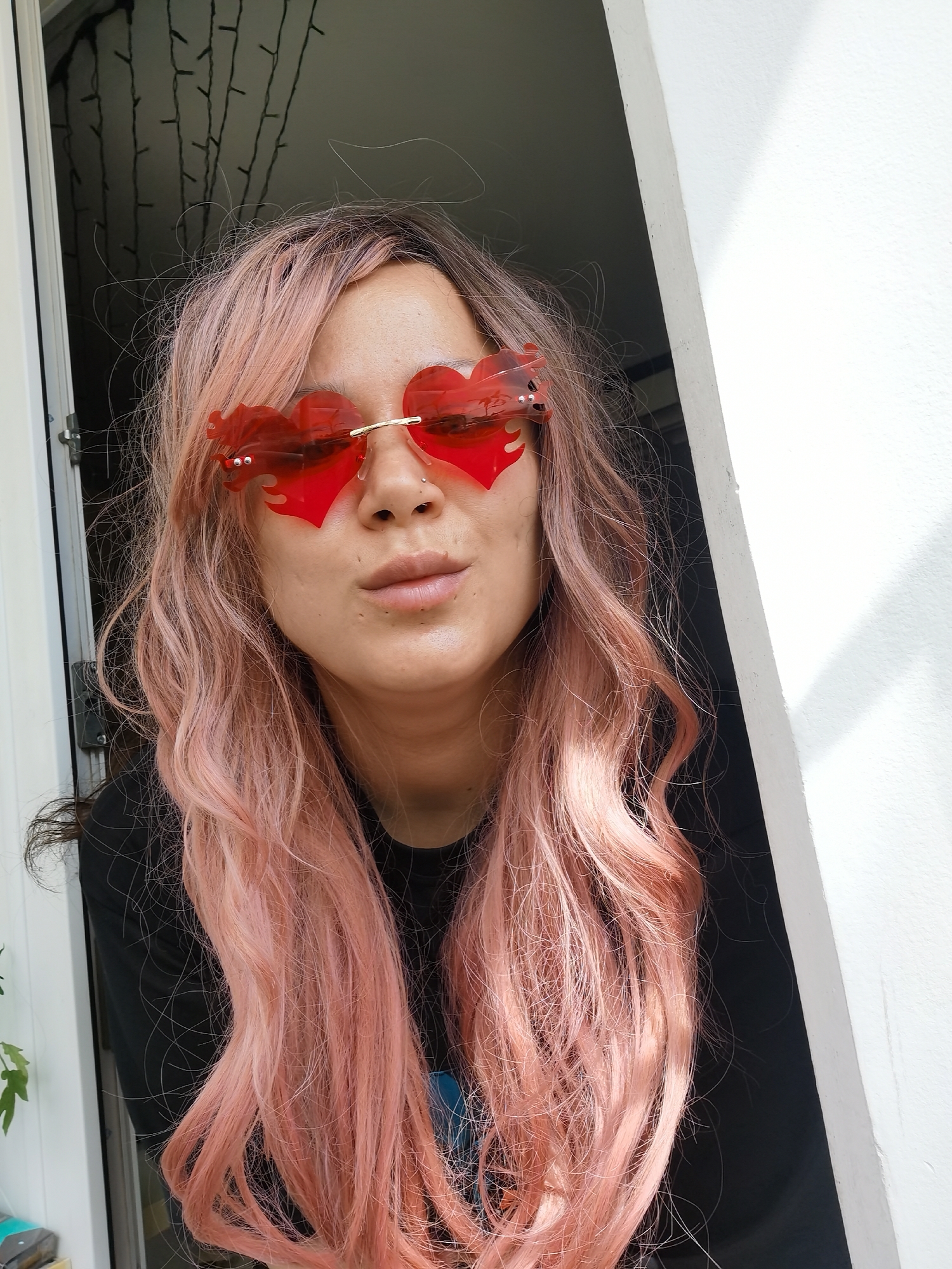 A woman with pink hair and heart-shaped red sunglasses stands indoors, likely near a doorway or window, as suggested by the surrounding white frame. She wears a black t-shirt and has a piercing in her nose. Behind her, through the window, there are draped lights. The location is Chaikovsky, Russia.