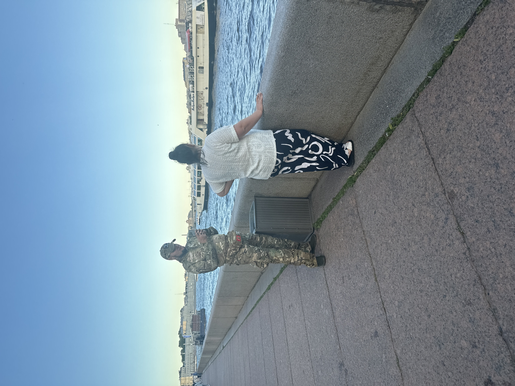 Two individuals are observed on a paved embankment adjacent to a body of water. On the left, a person dressed in camouflage military attire, including a cap, is looking at their mobile phone. They have a fanny pack with a red pouch around their waist. Next to this individual is a grey trash receptacle. To the right, another person is standing with their back to the camera, facing the water. They are wearing a white textured shirt and black and white patterned pants. They are leaning on the stone embankment, with their right hand resting on it. In the background, several boats and buildings are visible along the waterfront. The sky is clear and blue, indicating daylight. The scene appears to be set in Saint Petersburg, Russia, given the architectural style of the buildings and the presence of boats on what is likely the Neva River.